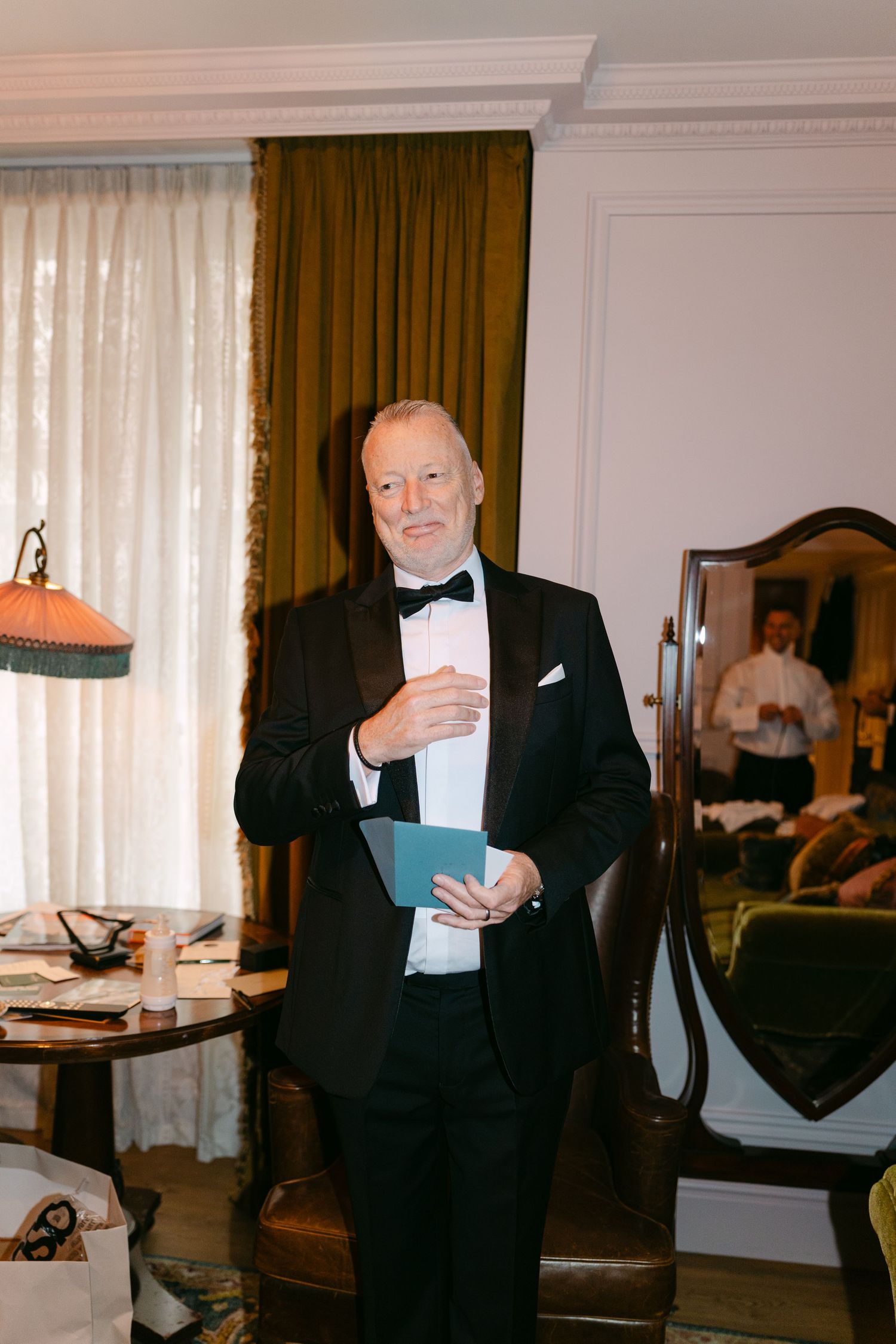A person in a black tuxedo holds papers while standing in an elegantly furnished room.
