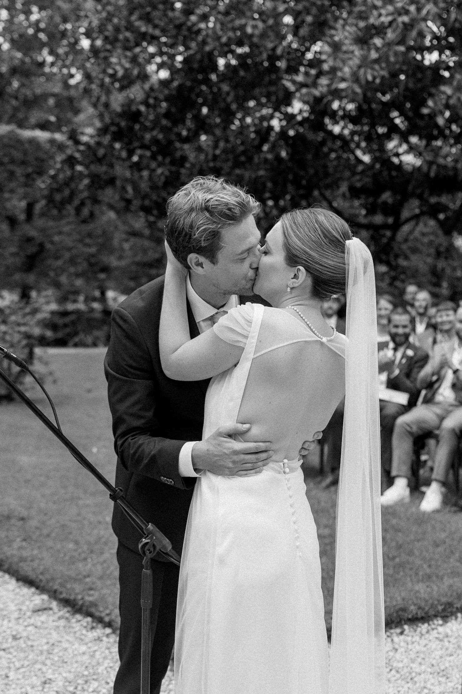 A romantic black and white wedding photo of a couple sharing their first kiss on a gravel path surrounded by greenery.
