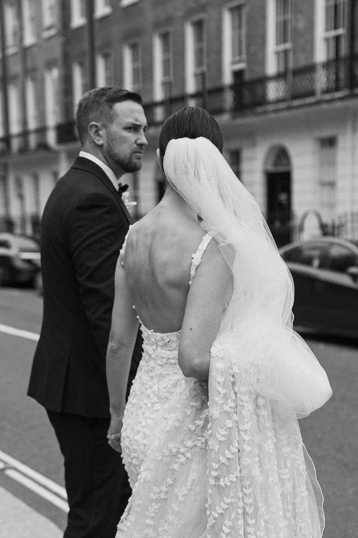 Bride in detailed lace dress with groom in formal suit on city street.