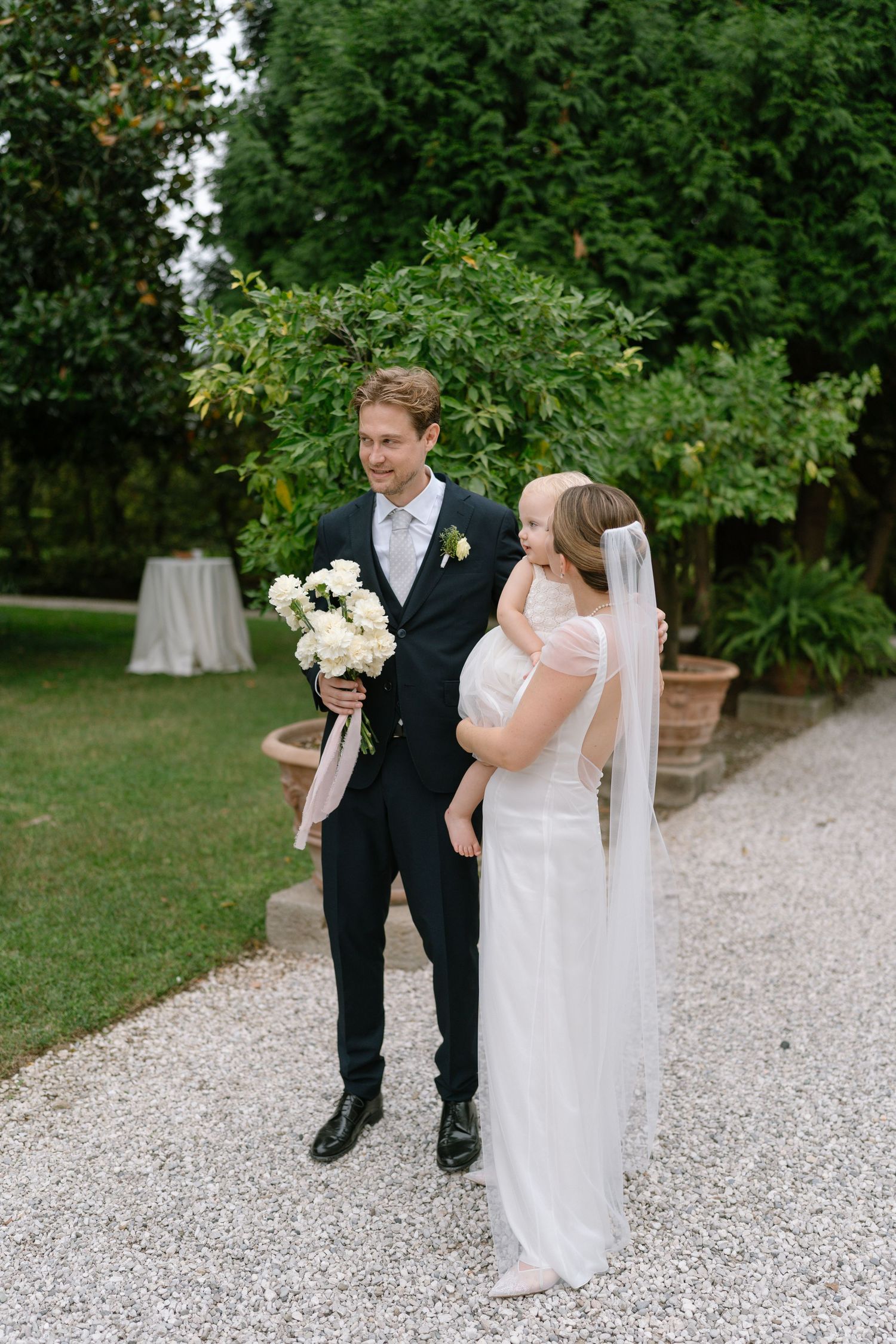 A couple walks along a white gravel path surrounded by lush greenery during their wedding day.