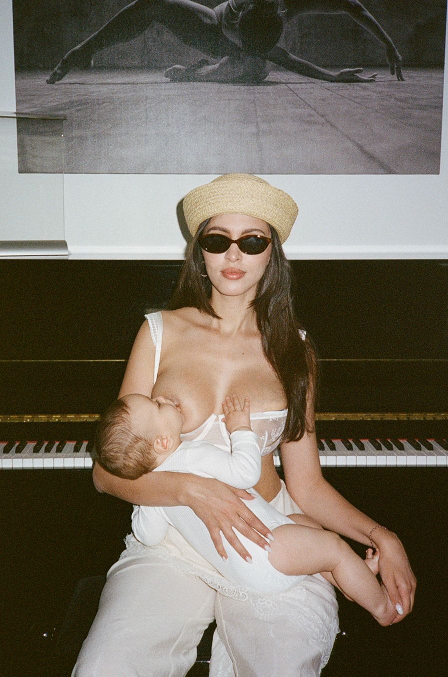 Woman in white outfit and straw hat breastfeeding baby while sitting in front of a piano.