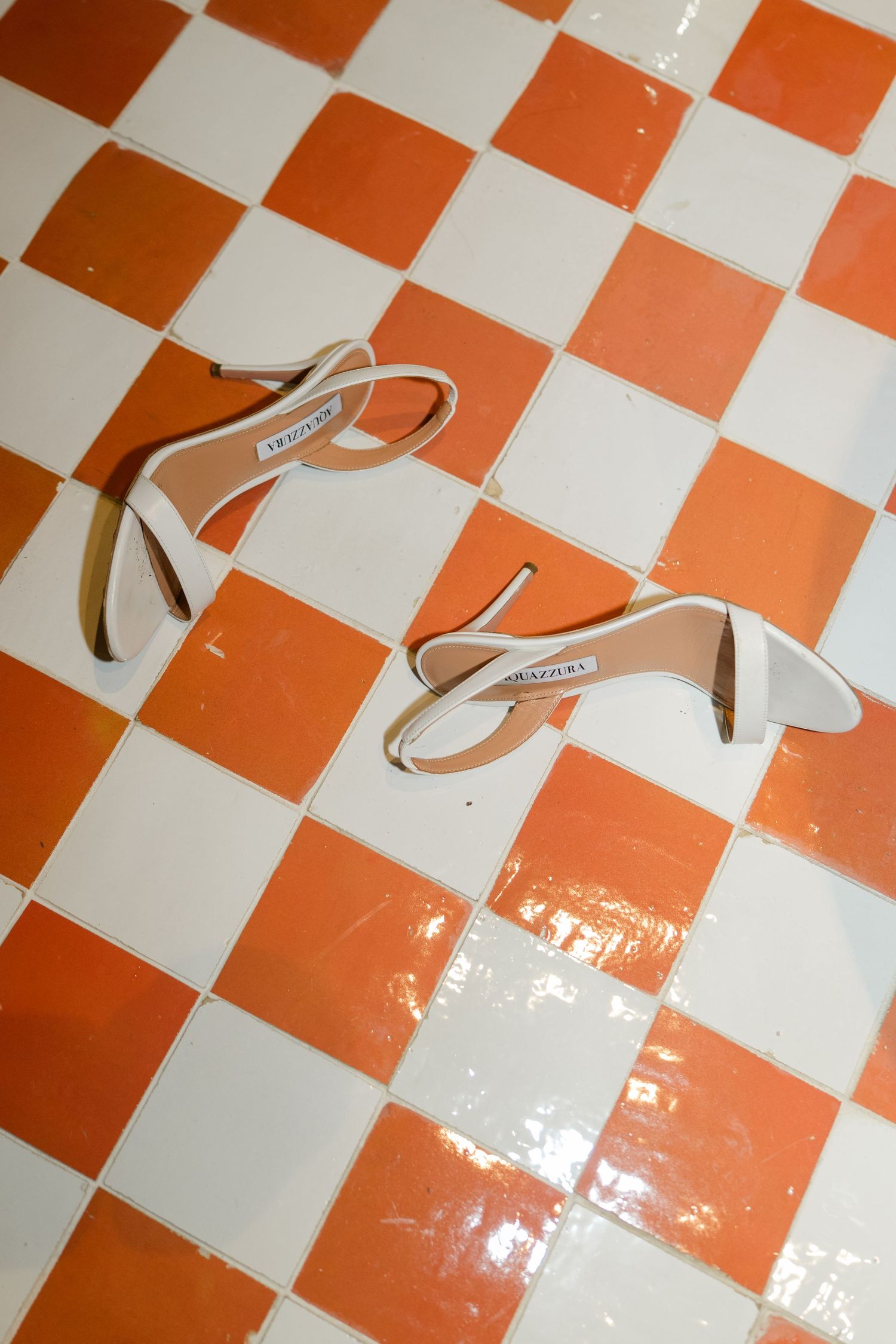 White strappy heeled sandals placed on glossy orange and white checkered tile floor.