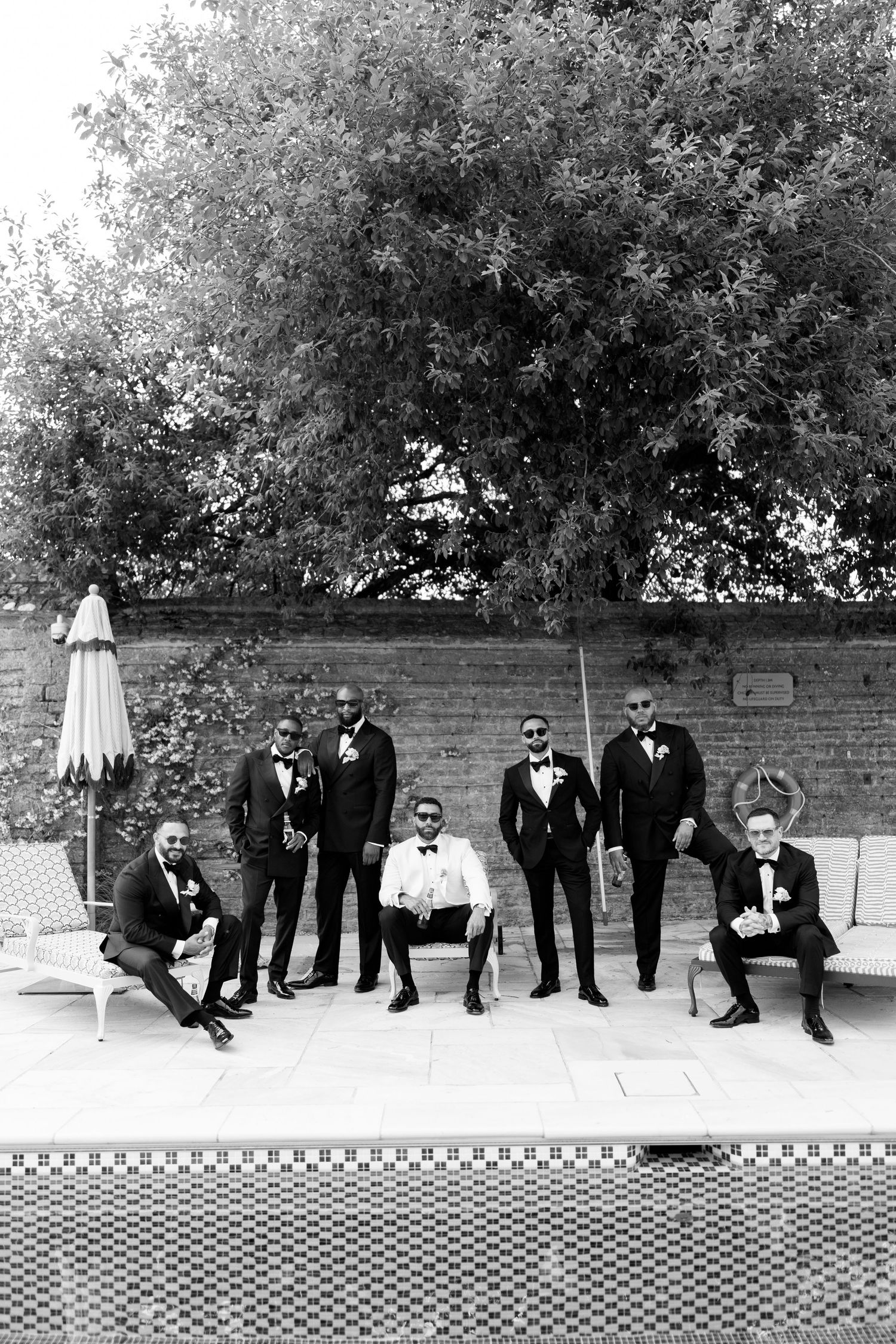 Groom and groomsmen in black tuxedos pose poolside in stylish black and white photo.