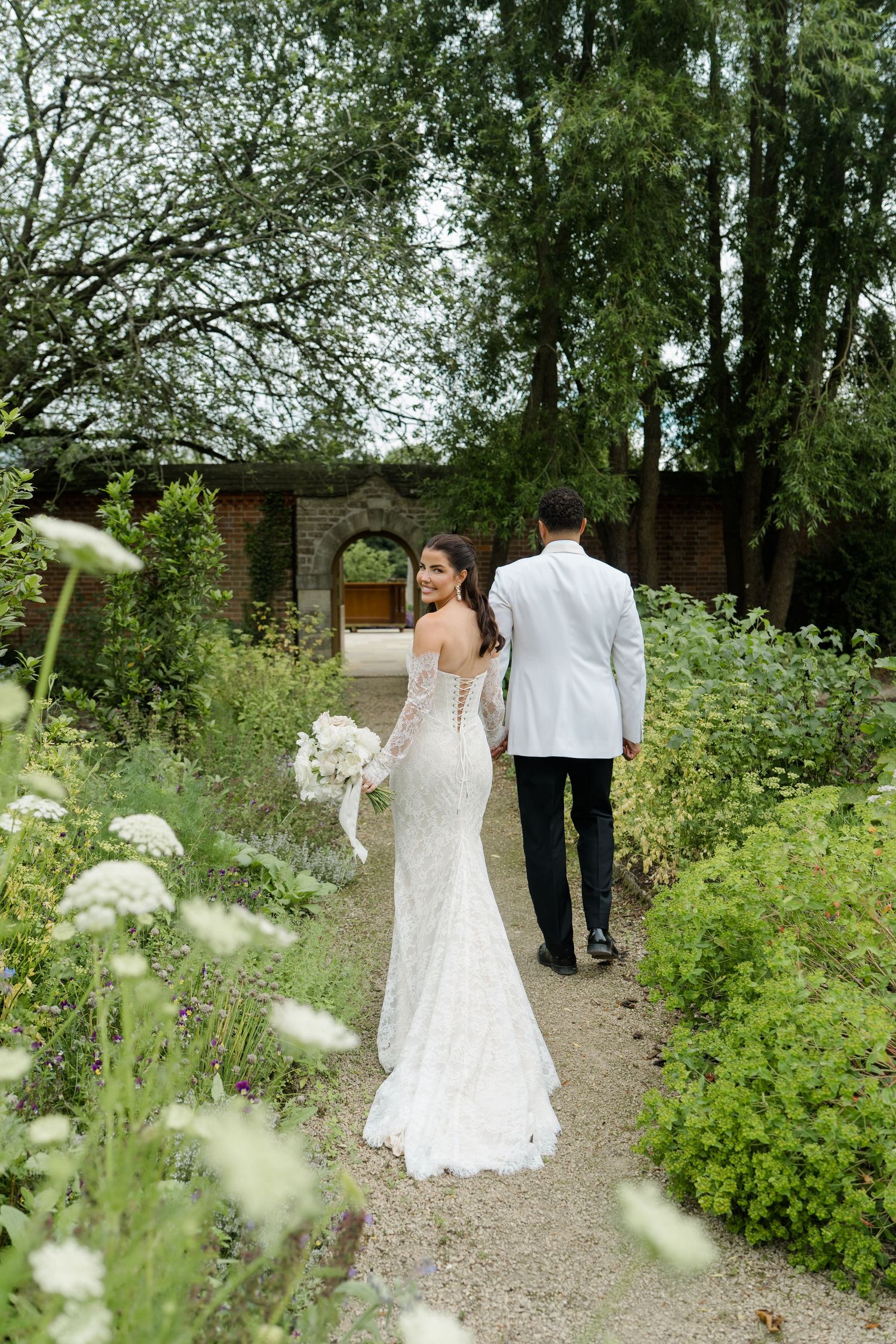 Bride in lace off-shoulder gown holds white bouquet while walking with groom through lush garden path.