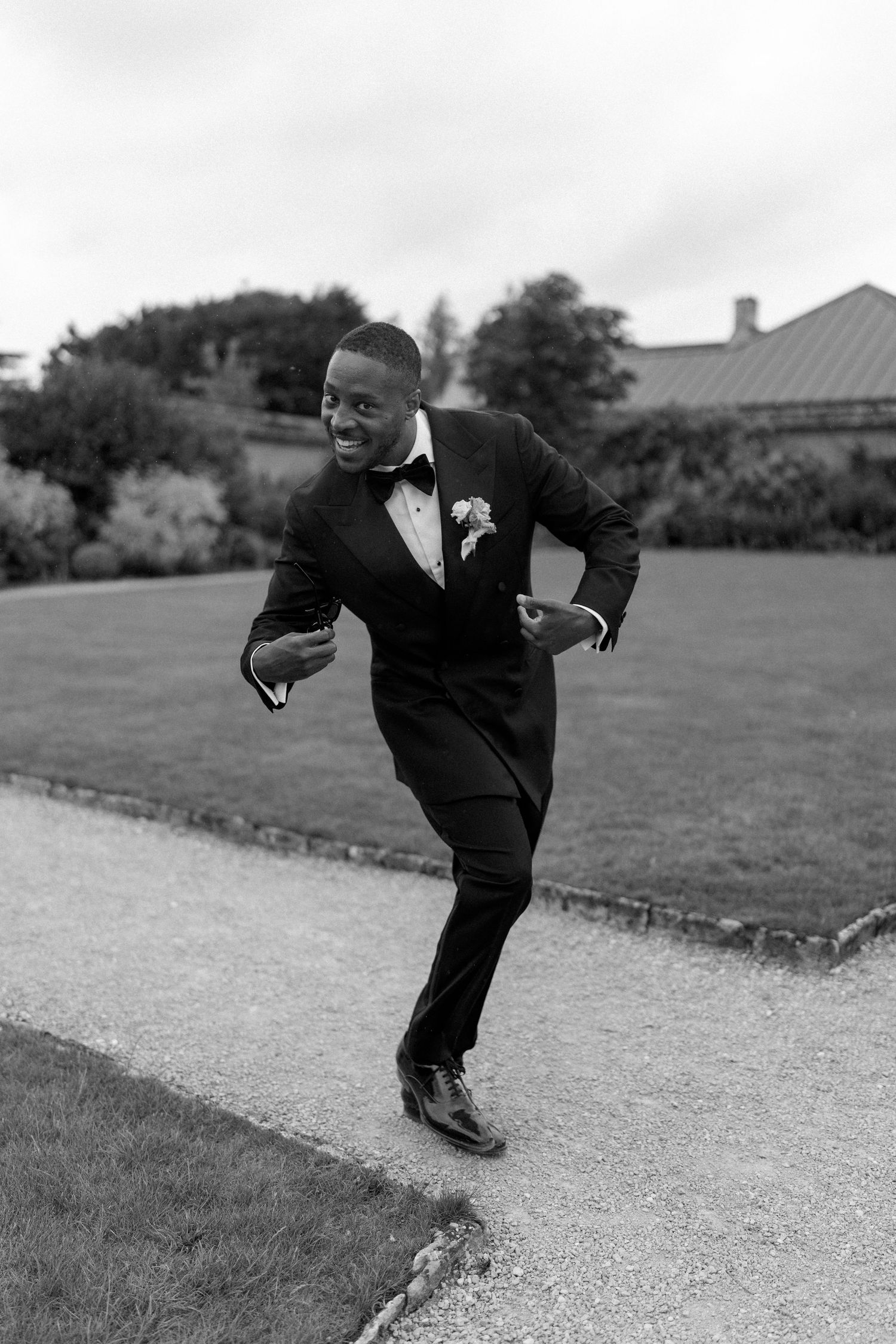 Joyful groom in black tuxedo dancing playfully on a garden path in black and white photo.