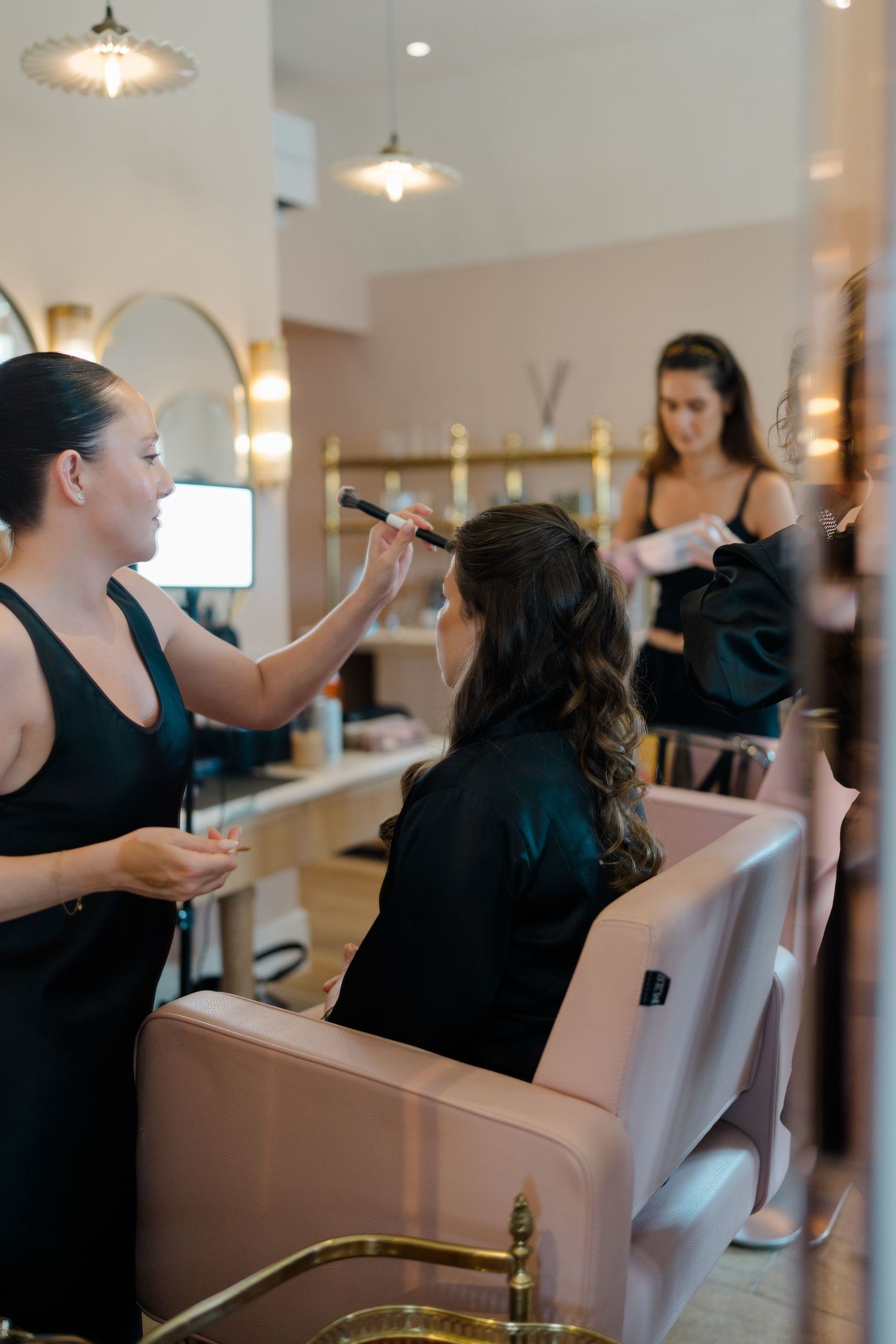 A makeup artist applies makeup to a bride in a pink salon chair while another woman gets ready in the background.