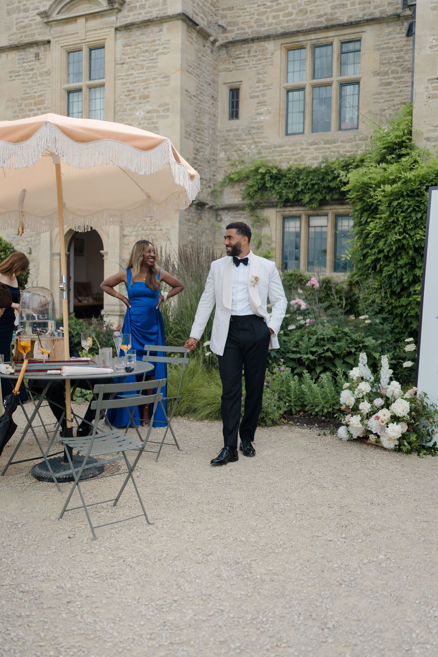 Groom in white tuxedo jacket stands in garden courtyard of stone manor house with wedding guests nearby.