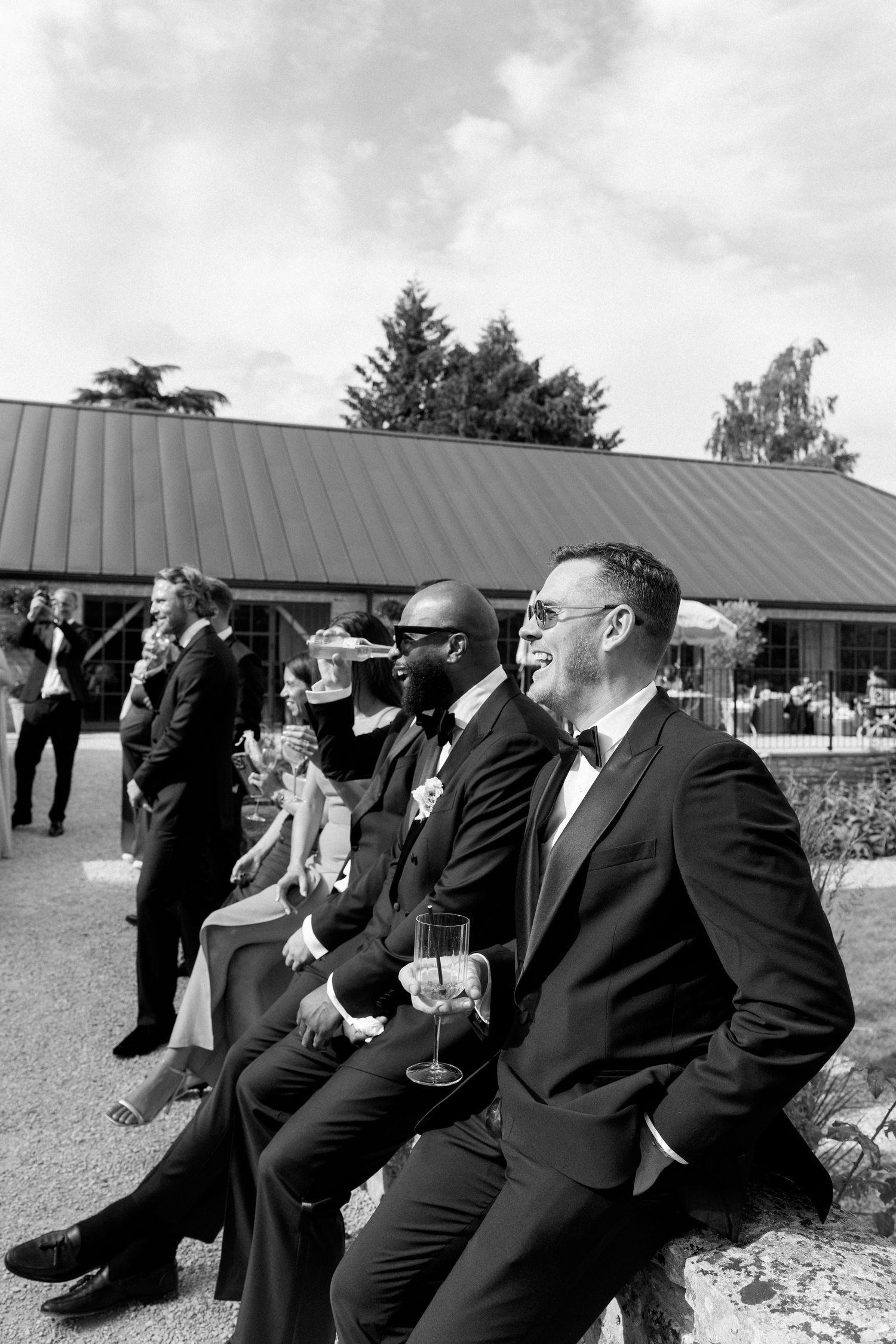 Groomsmen in black tuxedos relax on stone wall holding champagne glasses at outdoor wedding reception.