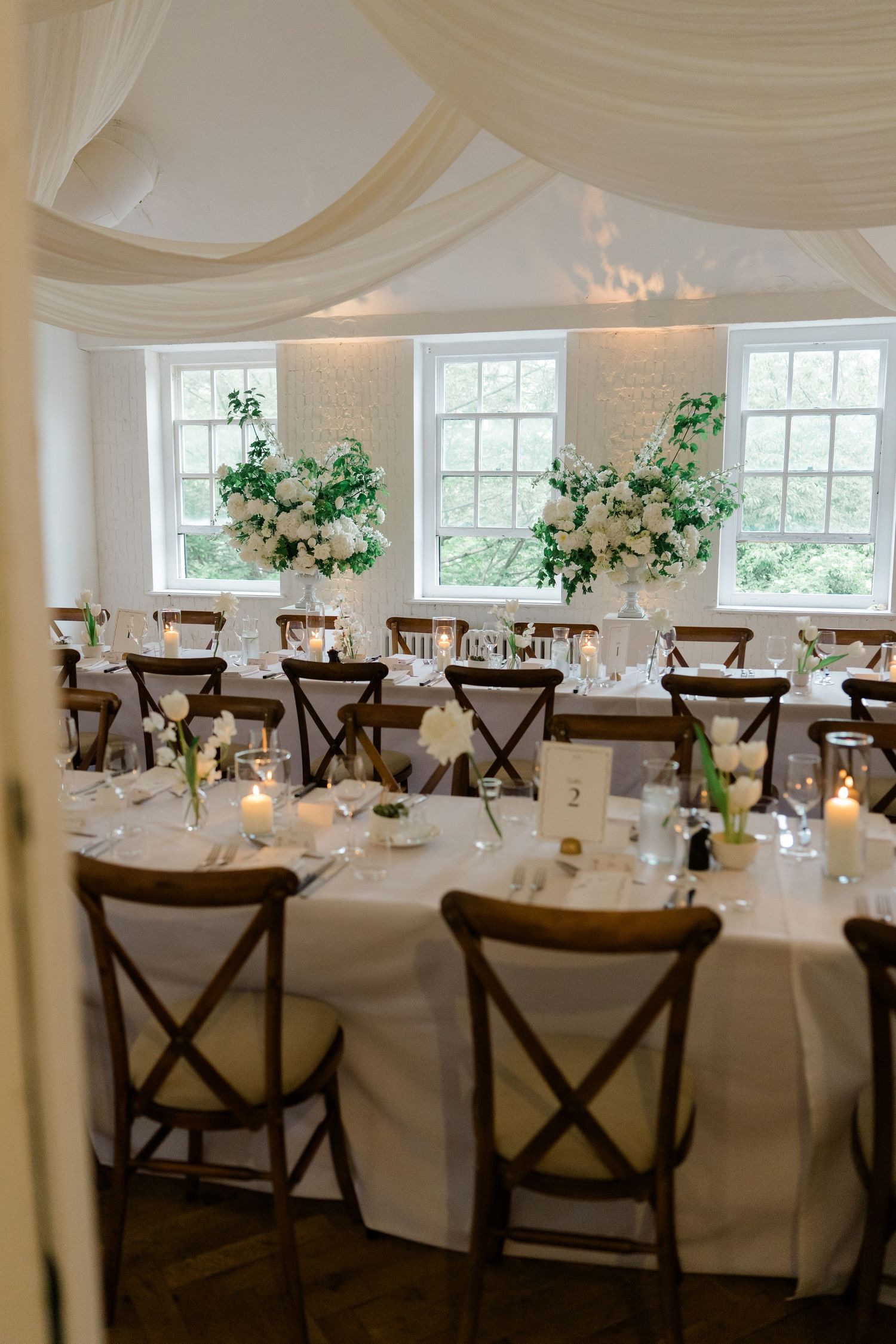 Elegant wedding reception room with round tables, wooden chairs, white draping, and tall floral arrangements.