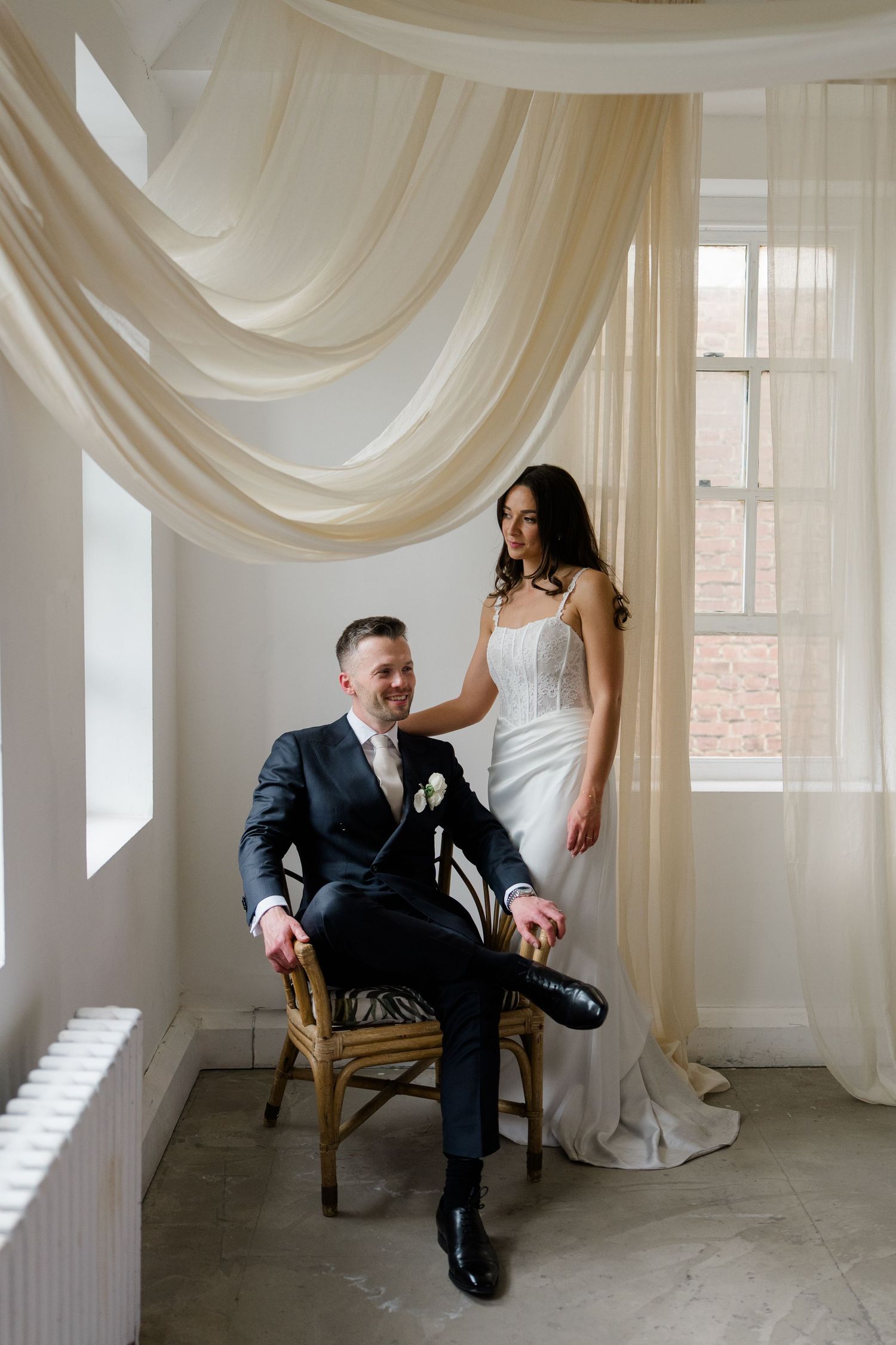 Groom sits in wicker chair while bride stands beside him under flowing cream drapes by window.