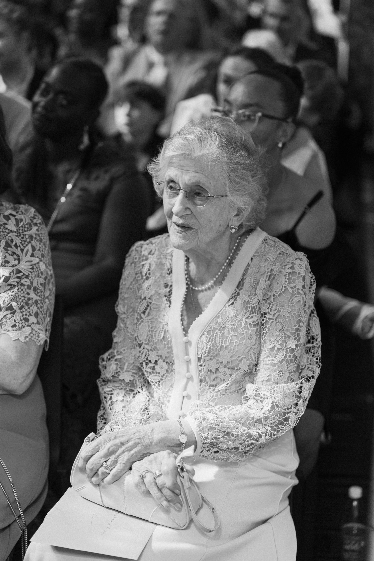 Elderly wedding guest in lace jacket and pearl necklace seated at ceremony, smiling warmly.