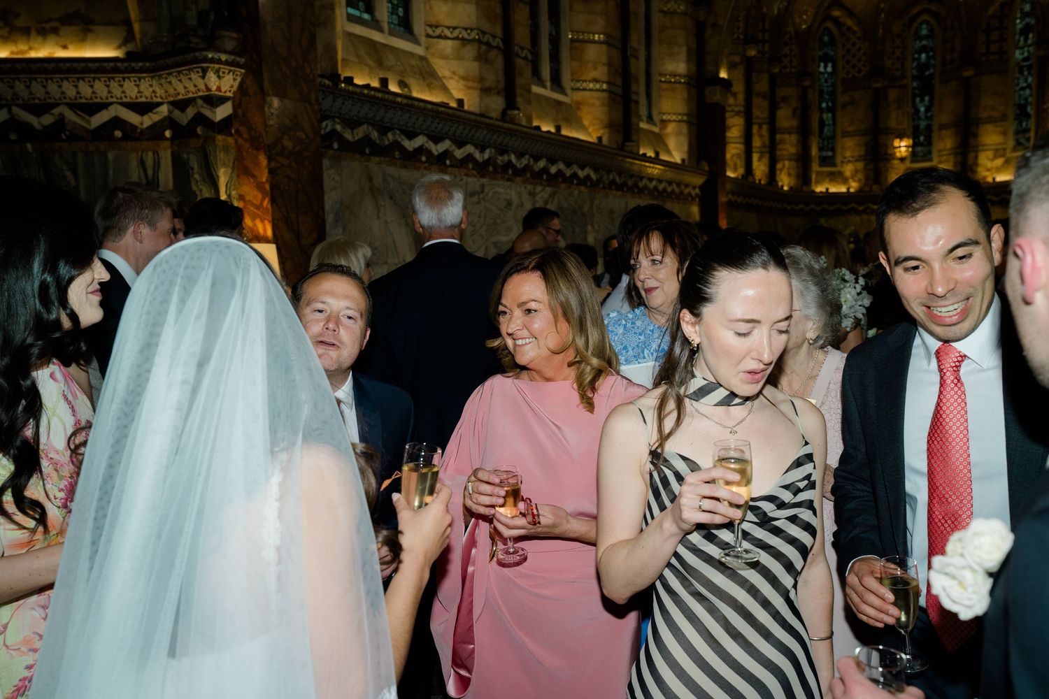 Wedding guests mingling and holding champagne glasses in an ornate historic venue during a cocktail reception.
