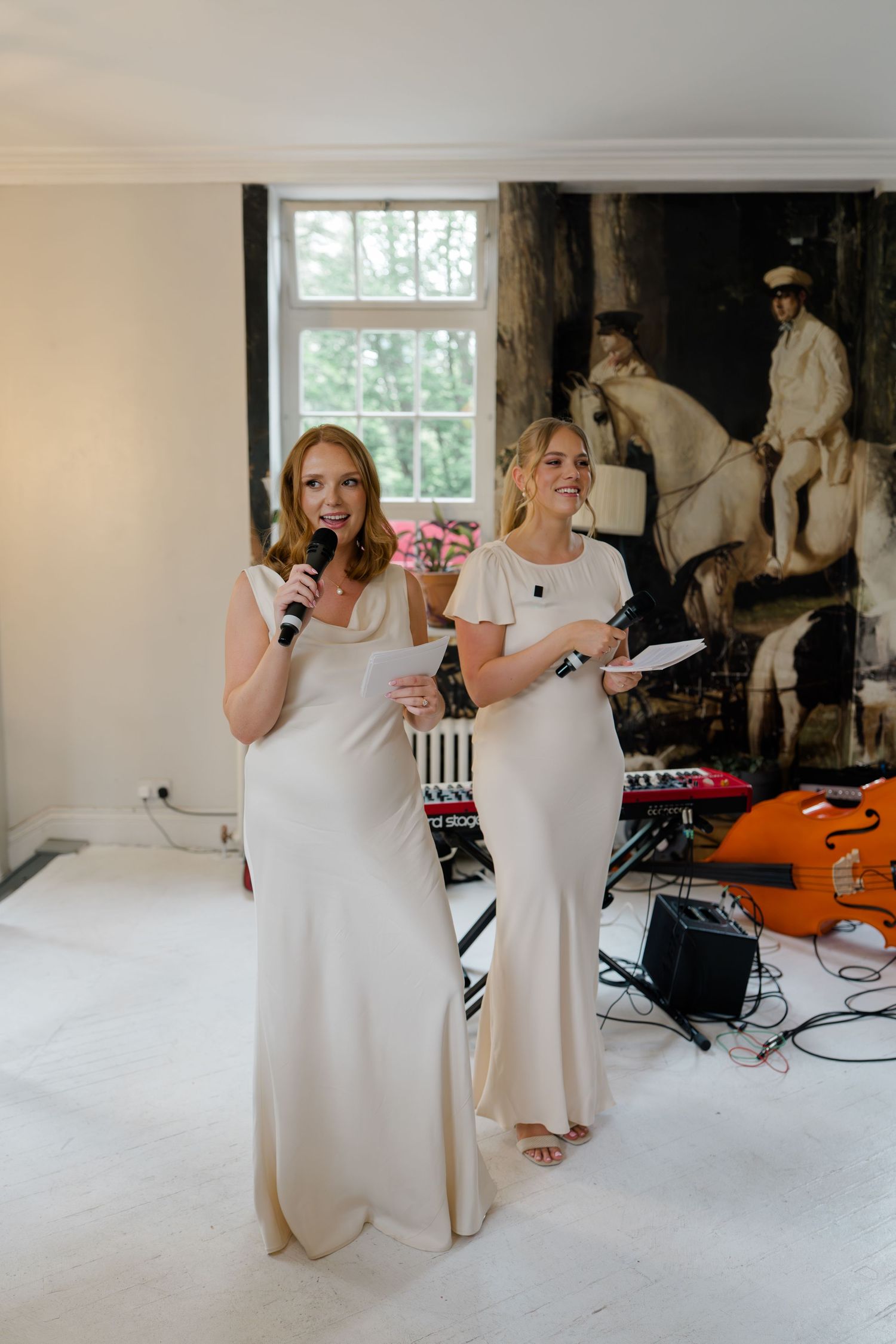 Two bridesmaids in ivory gowns give speeches at a wedding reception with musical instruments behind.