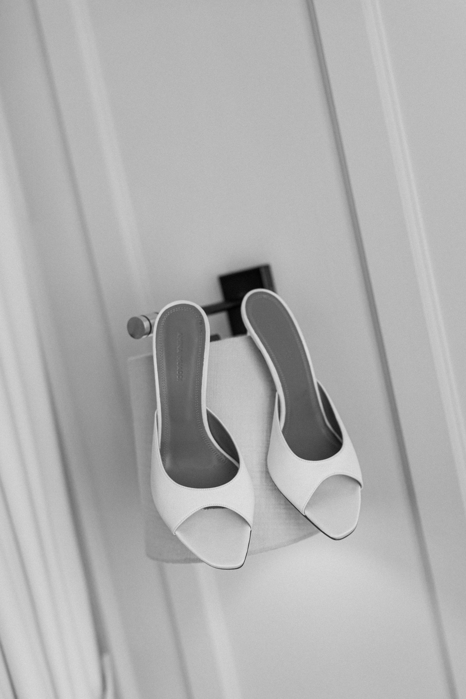 Black and white photo of elegant white open-toe bridal mule heels hanging on a door hook.