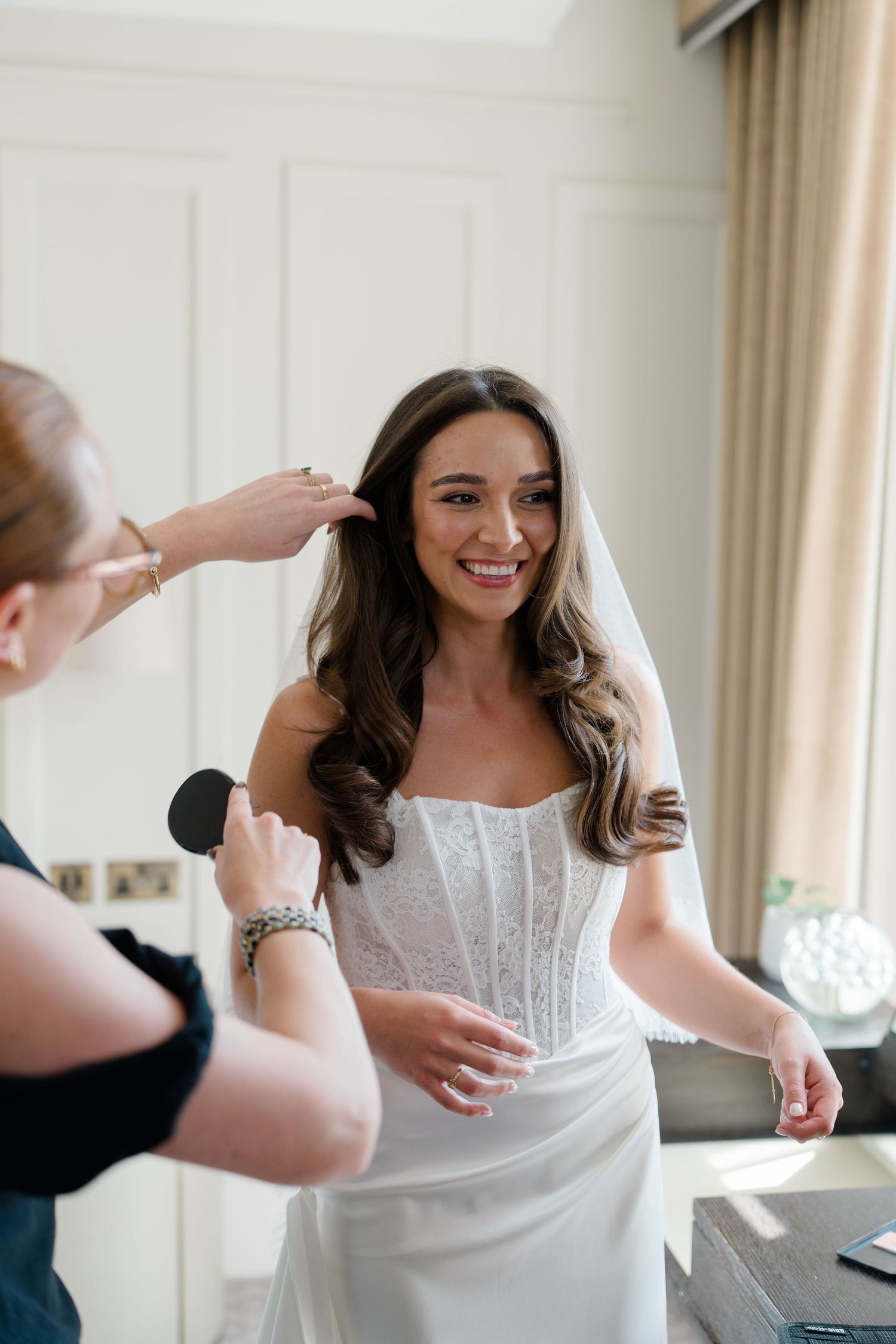Smiling bride in a white corset wedding dress getting her veil placed by a stylist in a bright room.