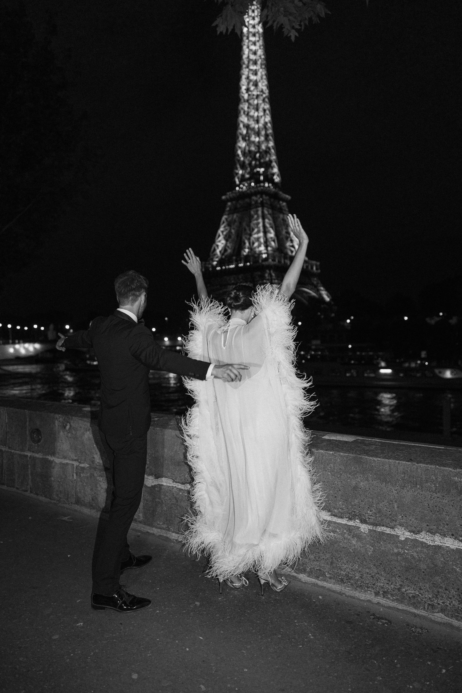 Black and white photo of a couple dancing by the Eiffel Tower at night, bride in feathered white gown.