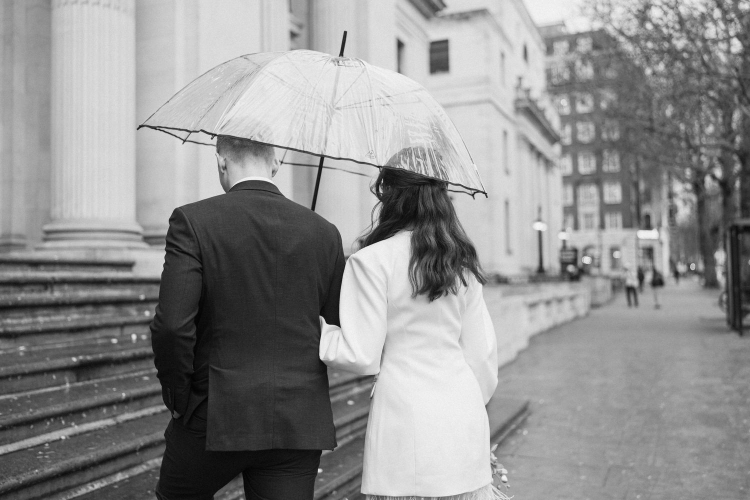 Bride and groom walk under clear umbrella past classical building columns in black and white.
