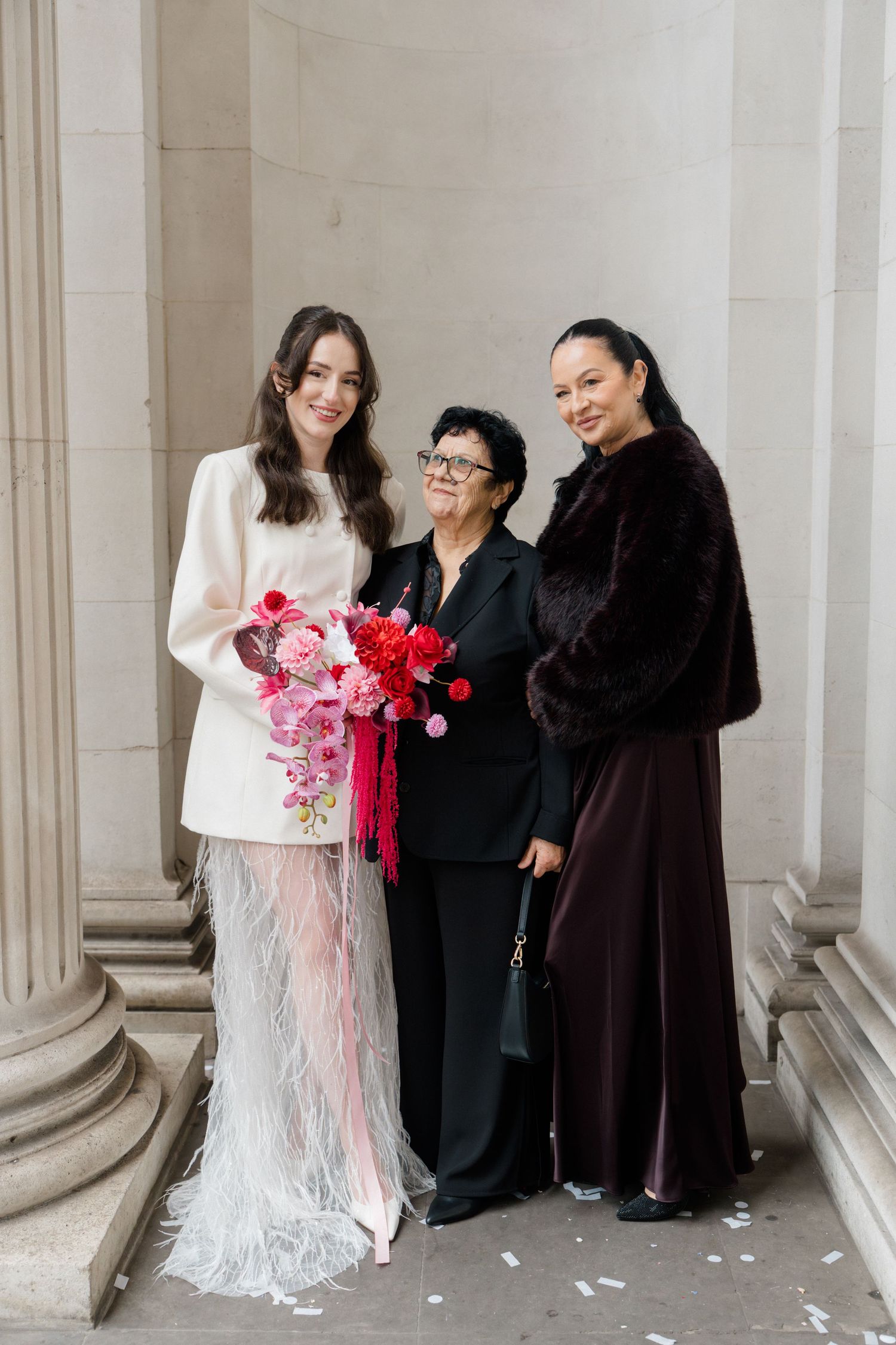 Bride in white mini dress with feathered skirt poses with two female guests holding pink red bouquet.
