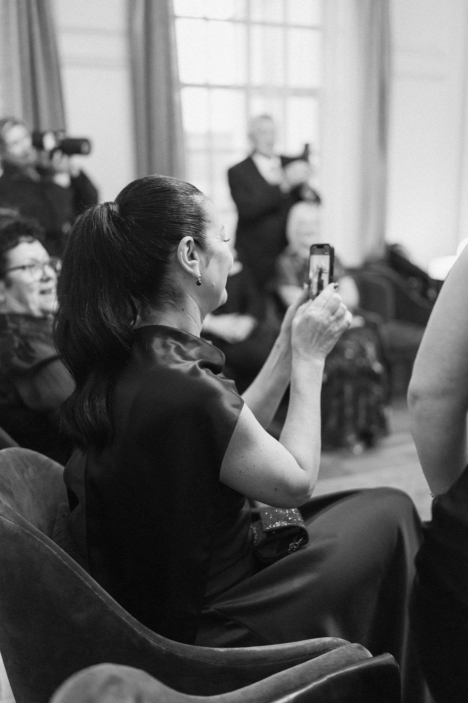 Black and white photo of woman in dark dress photographing a wedding ceremony on her smartphone.