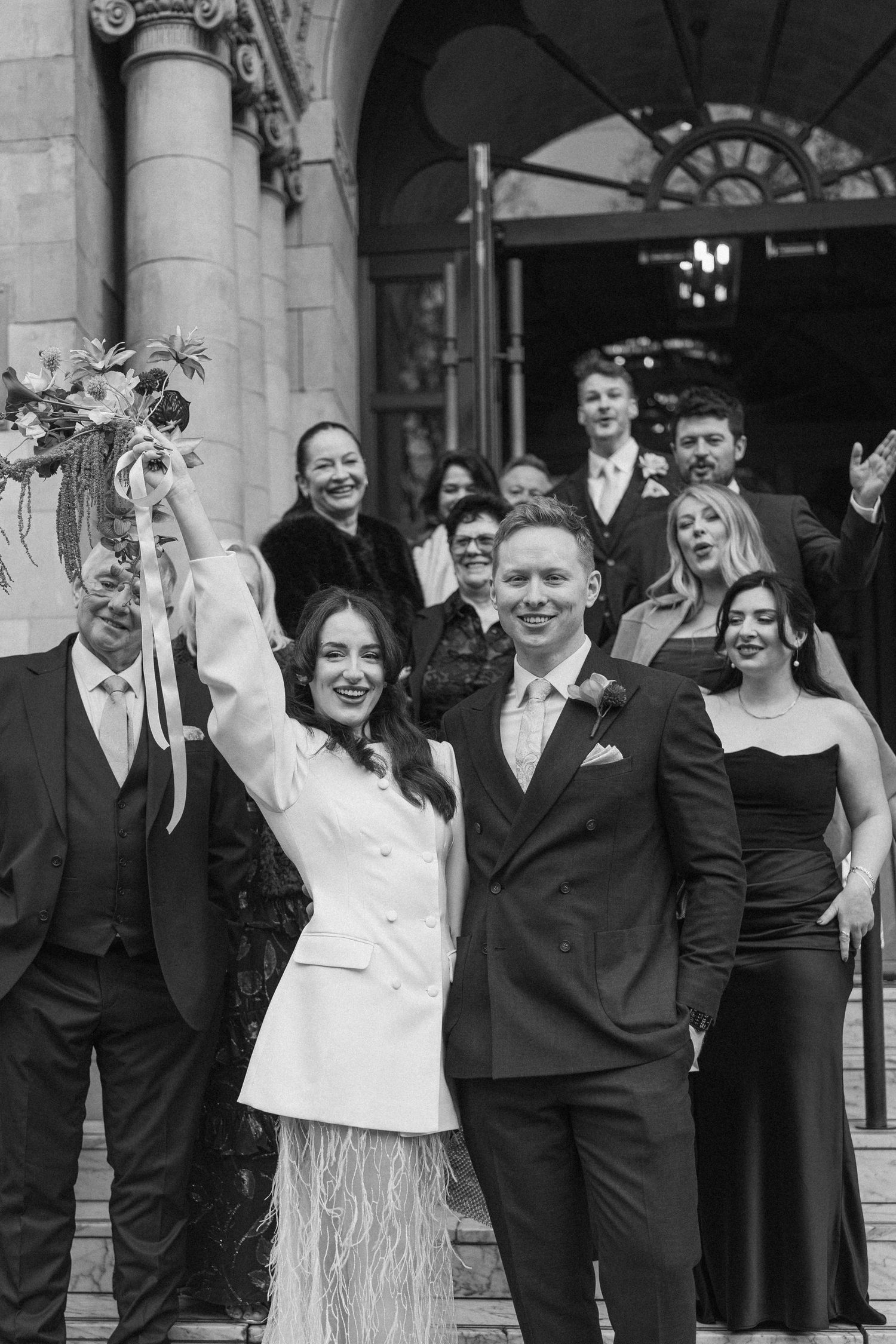 Black and white photo of joyful newlyweds celebrating with guests on grand building steps.