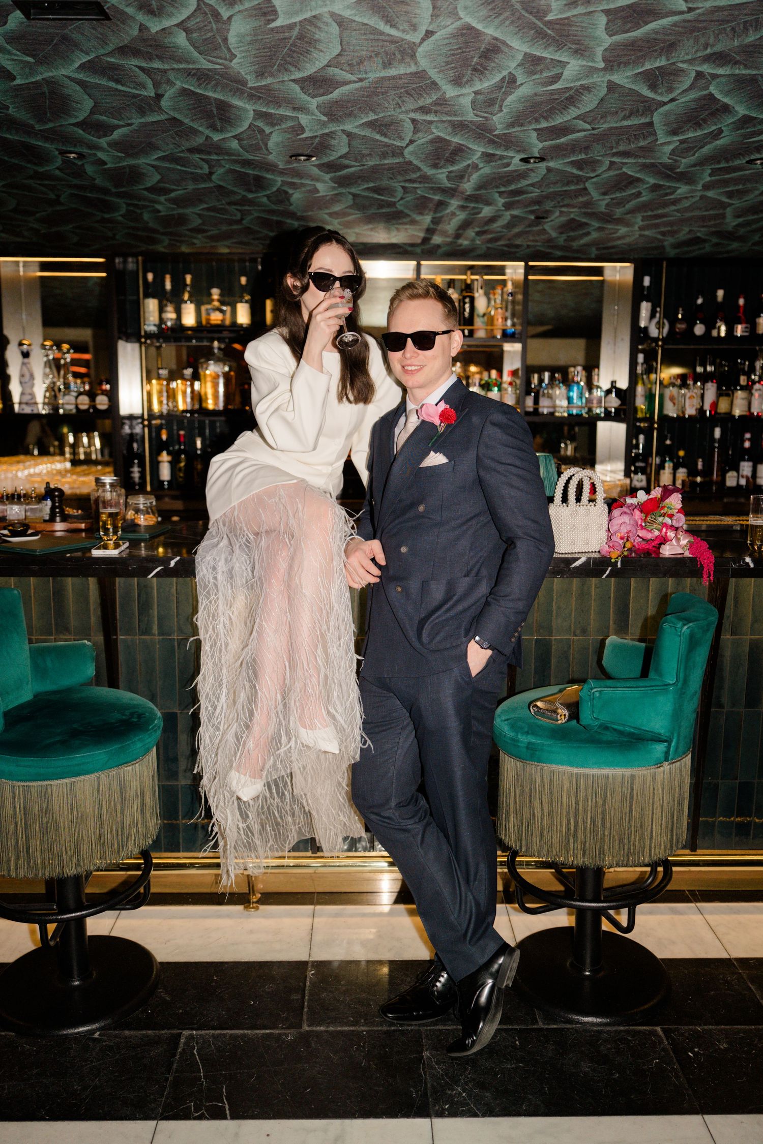 Stylish couple posing at a glamorous bar, bride in feathered skirt and groom in navy suit.