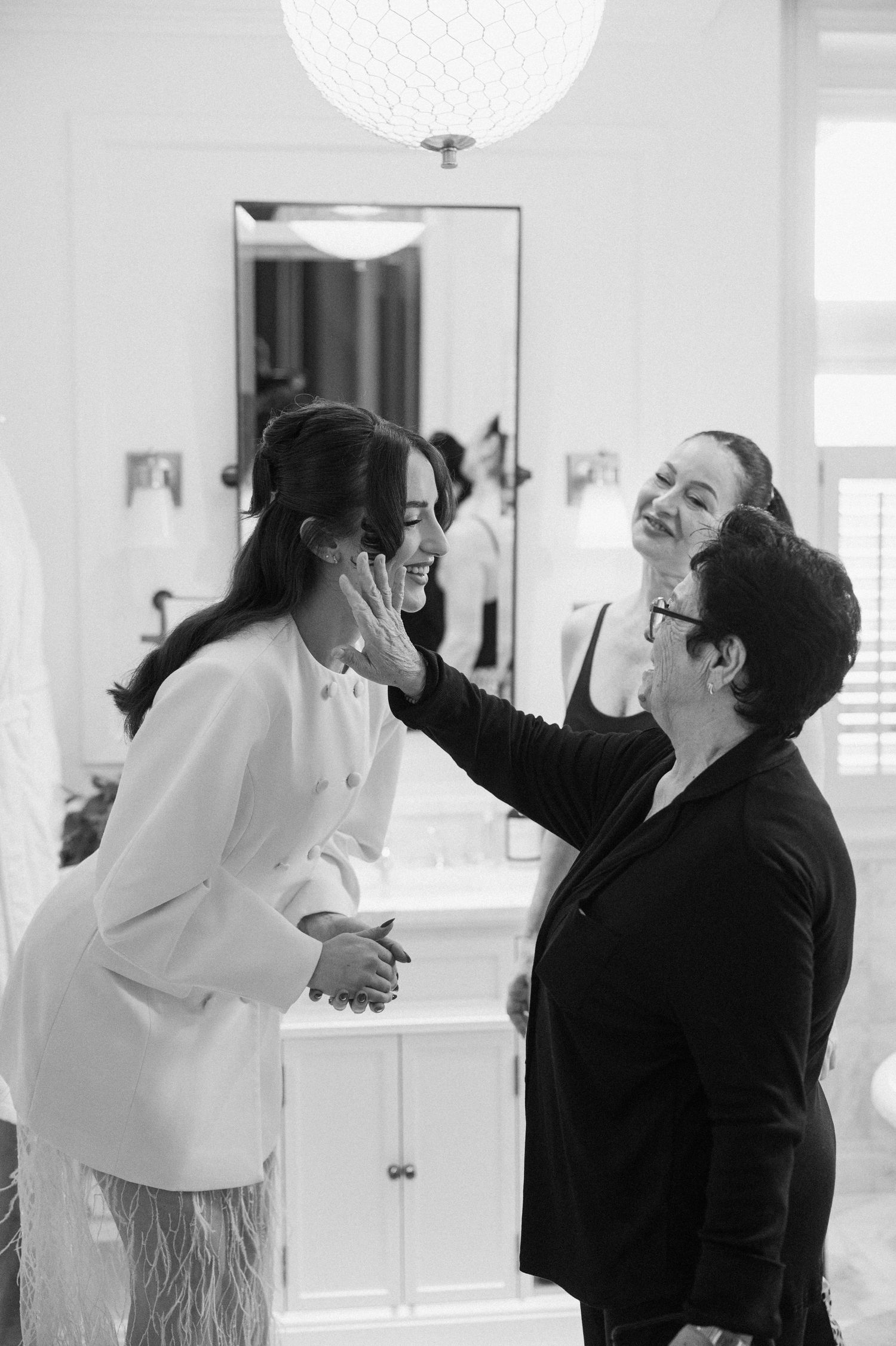 A black and white photo of a bride laughing as an older woman touches her face during wedding preparations.