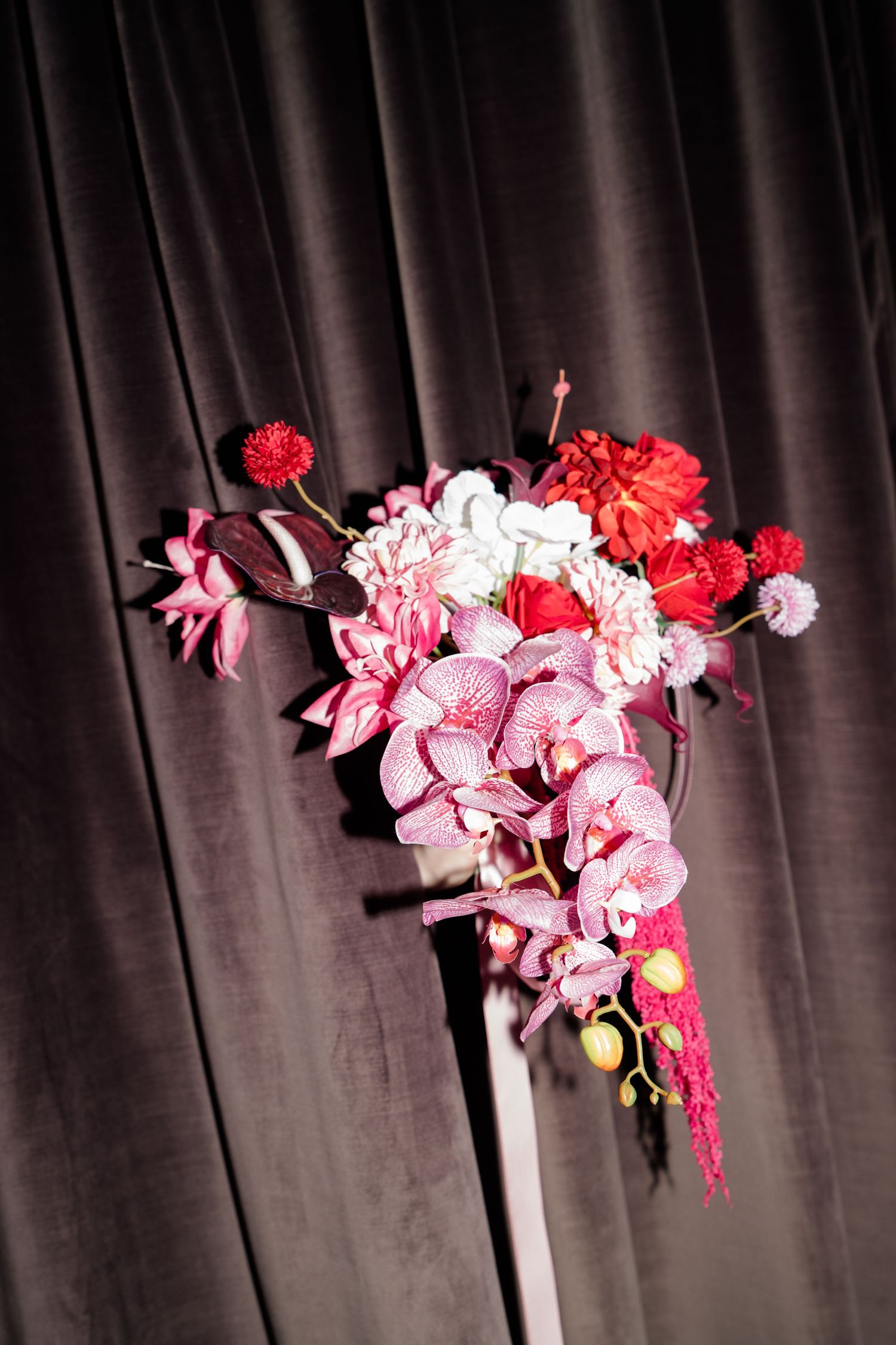 Stunning bridal bouquet featuring pink orchids, red dahlias and white flowers against dark curtain.