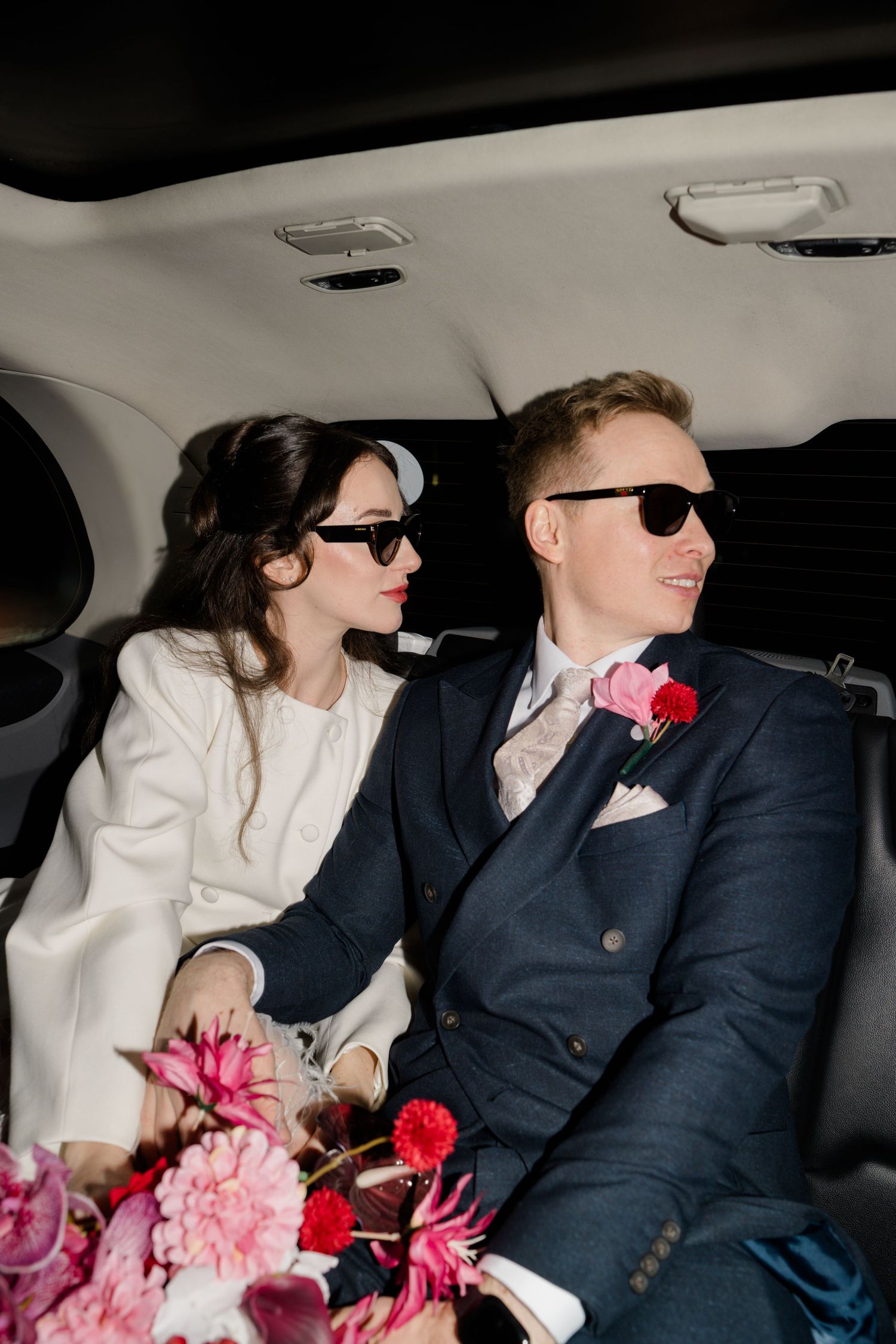 Stylish bride and groom wearing sunglasses sitting in luxury car with pink floral bouquet.