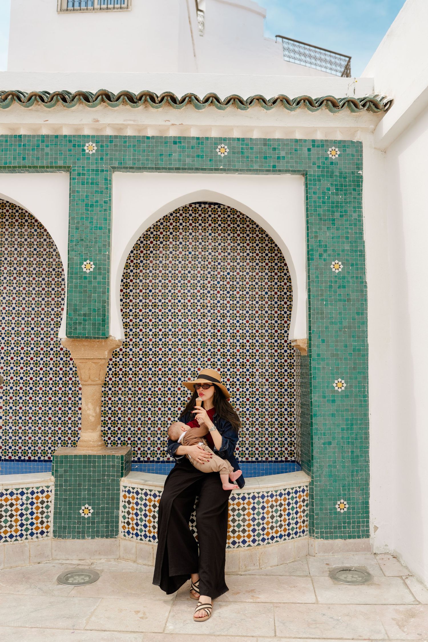Woman in straw hat holding baby in front of ornate Moroccan mosaic tile arches with green geometric patterns.