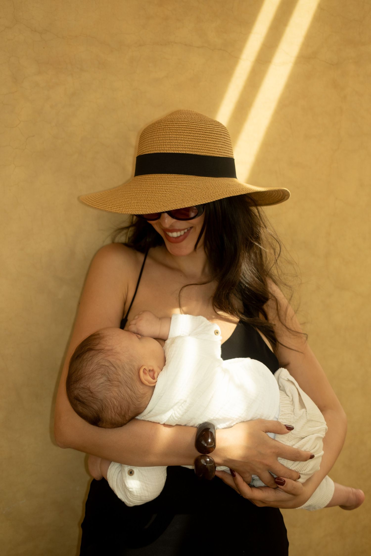 Smiling woman in wide-brim straw hat and sunglasses breastfeeding newborn baby in white wrap outdoors.