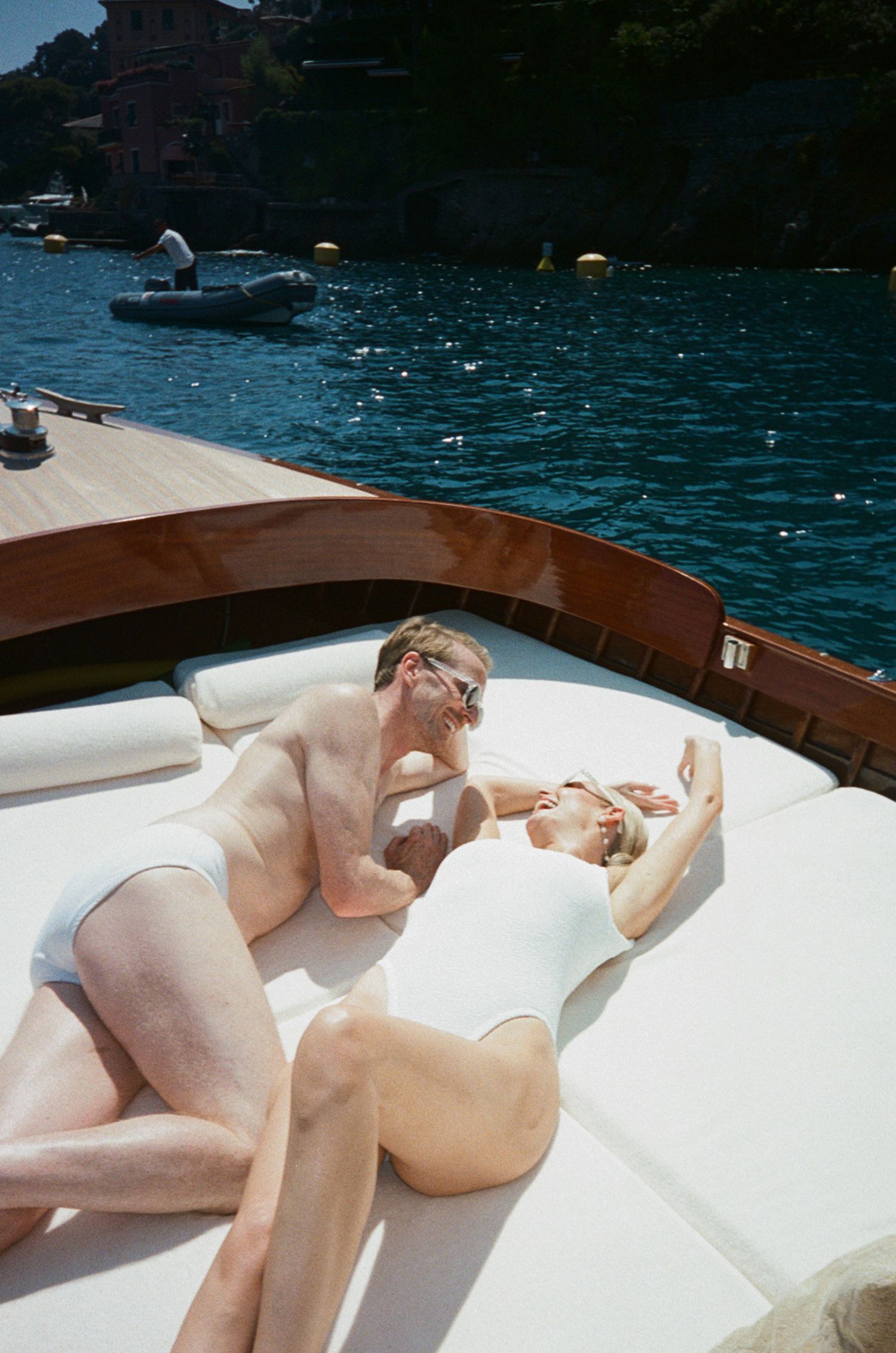 Couple in white swimwear relaxes together on luxury boat bow with turquoise Italian harbor water behind.