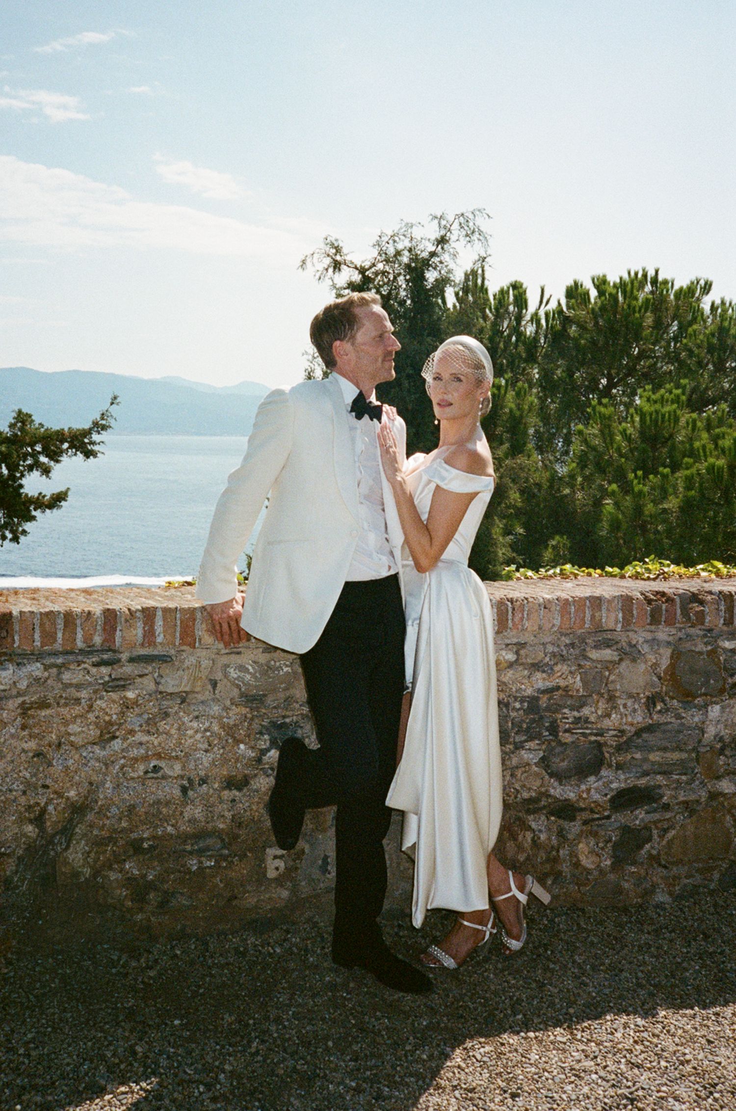 Bride and groom embracing by stone wall with stunning Mediterranean sea and mountains in background.