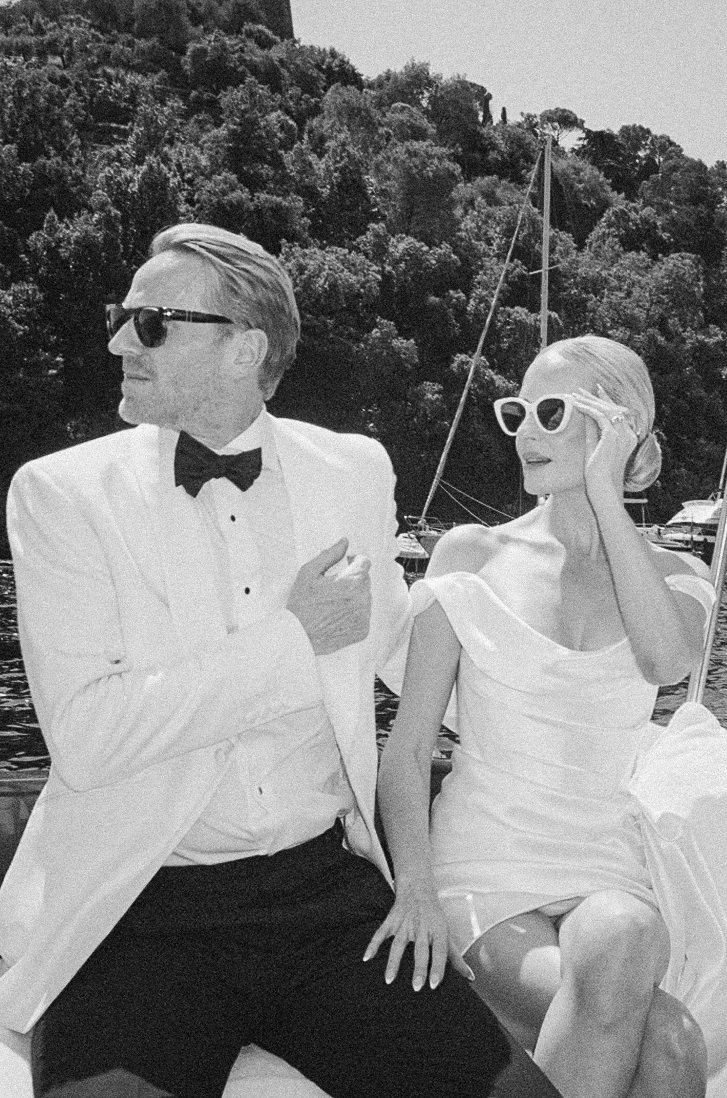 Elegant couple in white wedding attire on boat with sunglasses, black and white photo.