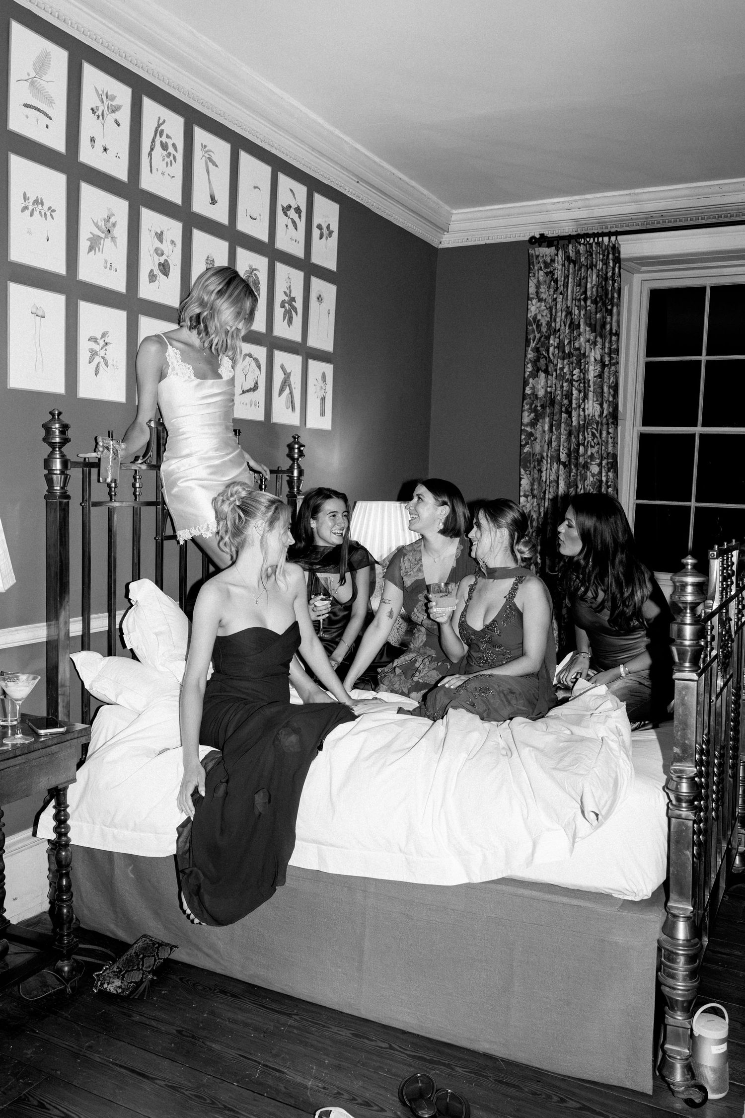 Black and white photo of bride standing on bed surrounded by laughing bridesmaids in a decorated bedroom.