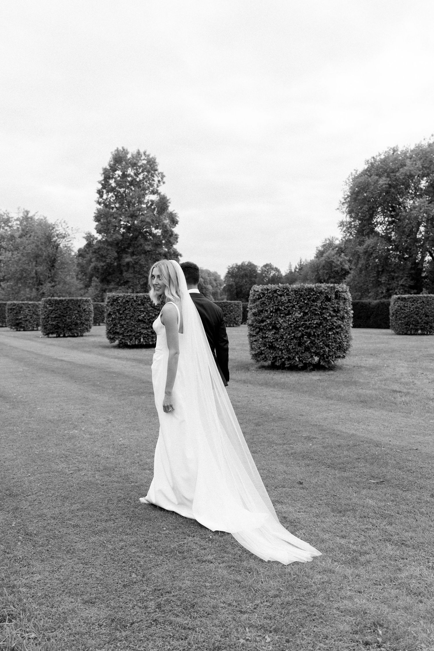 Bride in white gown and veil walks with groom through manicured garden hedges in black and white wedding photo.