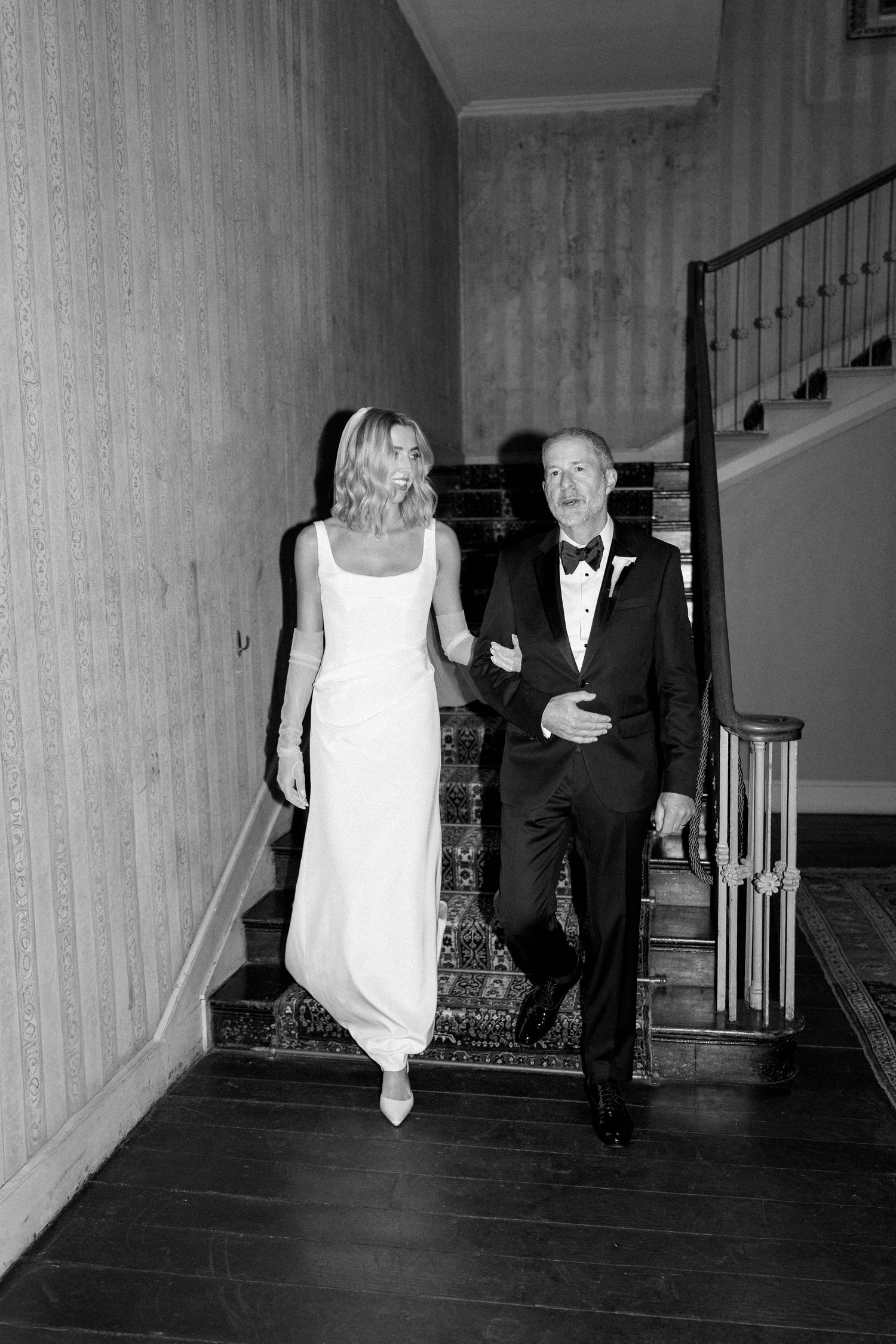 Bride in white gown walks arm-in-arm with man in tuxedo down elegant staircase in black and white photo.