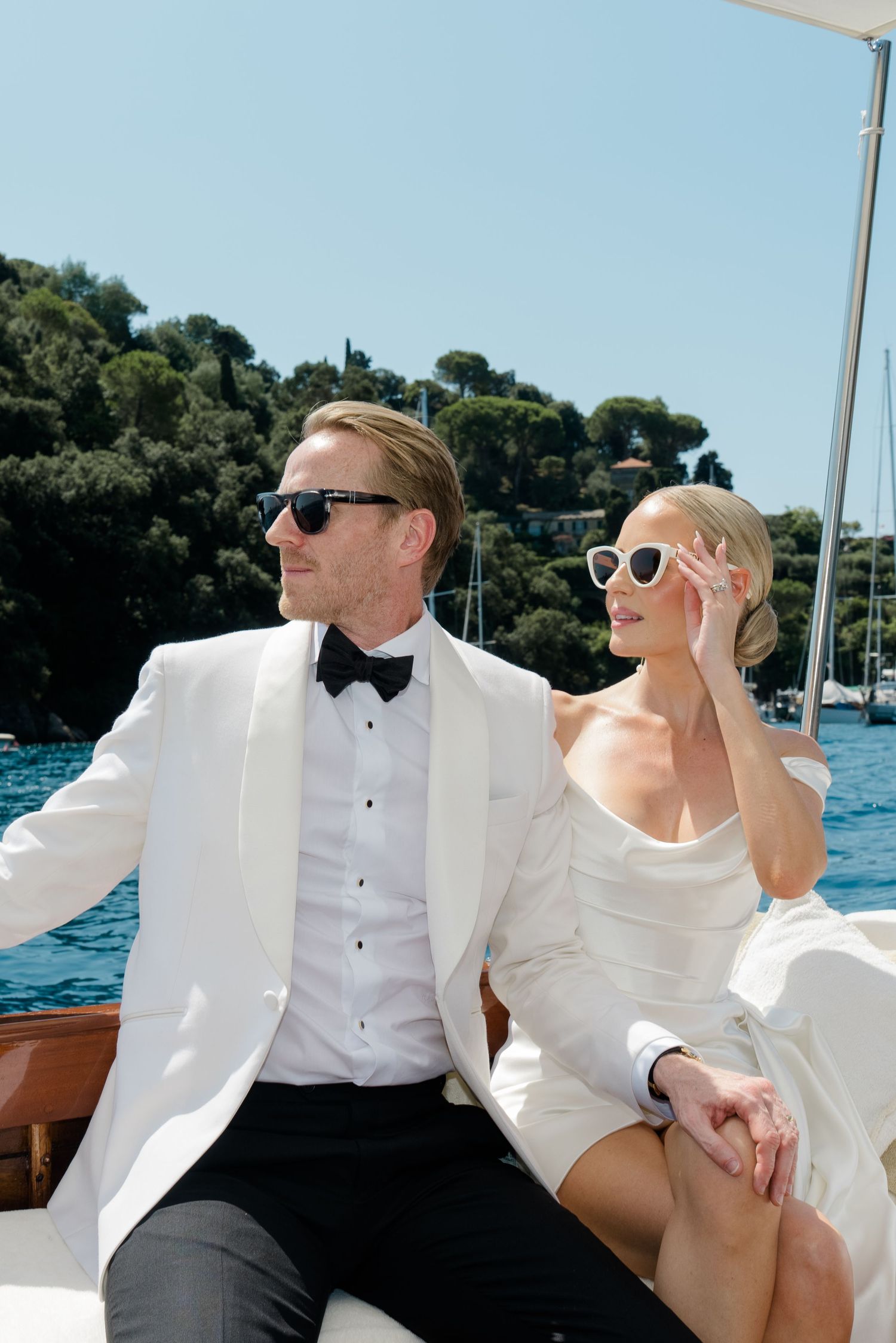 Stylish newlyweds in white wedding attire wearing sunglasses relaxing on a luxury boat in Italy.