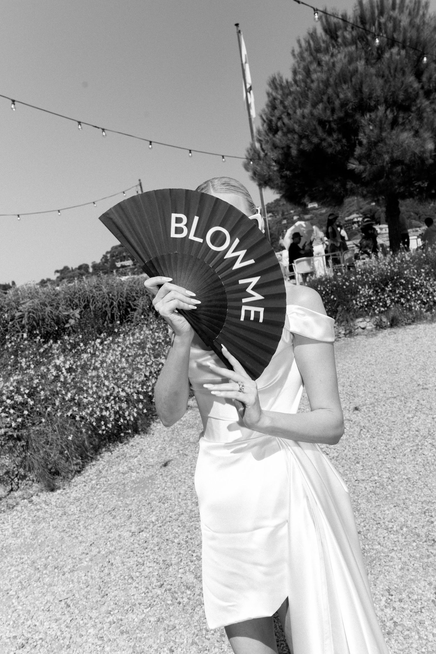 Bride in white dress holding a black hand fan reading Blow Me at an outdoor wedding reception.