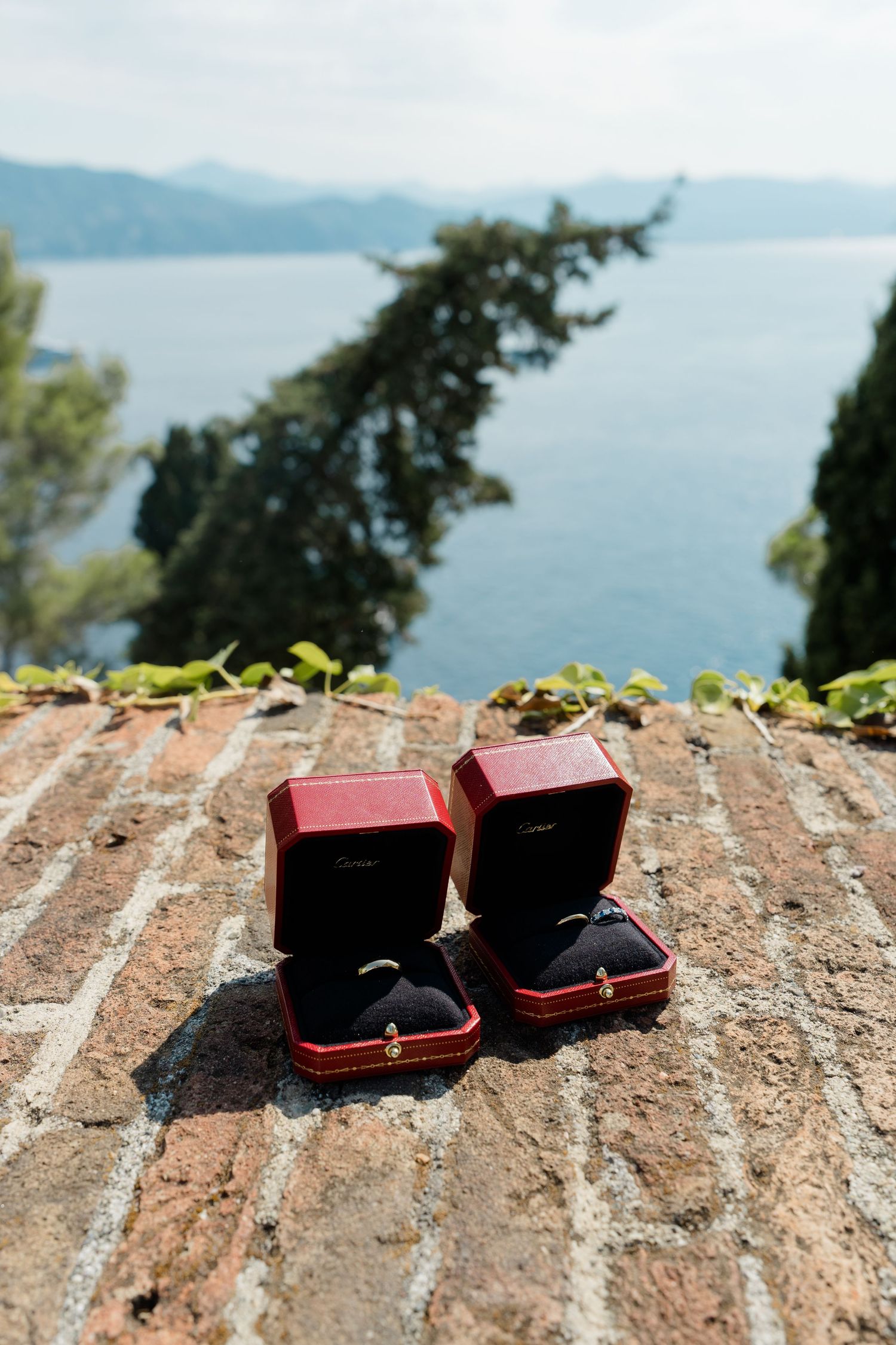 Two red Cartier ring boxes displayed on a rustic stone wall with a scenic blue sea backdrop.