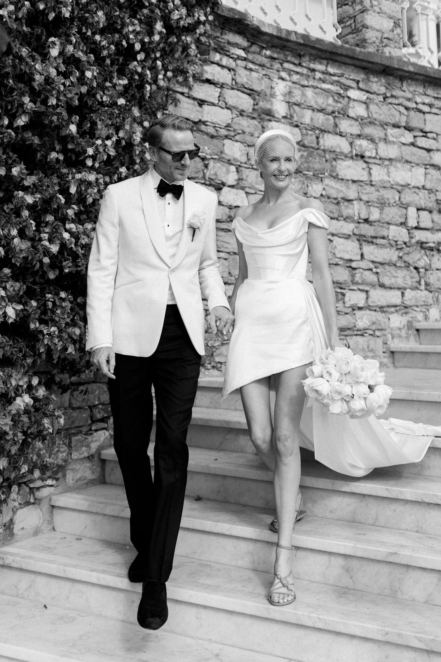 Elegant bride and groom descend stone steps, she in a short white dress holding roses, he in a white tuxedo jacket.
