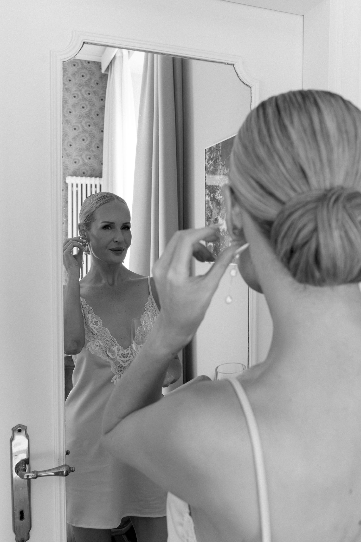 Black and white photo of a blonde bride putting on earrings while looking at her reflection in a mirror.