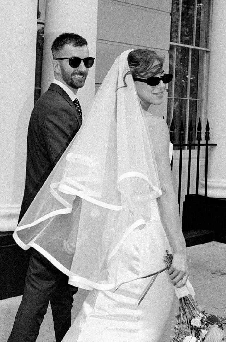 Black and white photo of a bride in veil and sunglasses holding bouquet with suited groom outside a building.