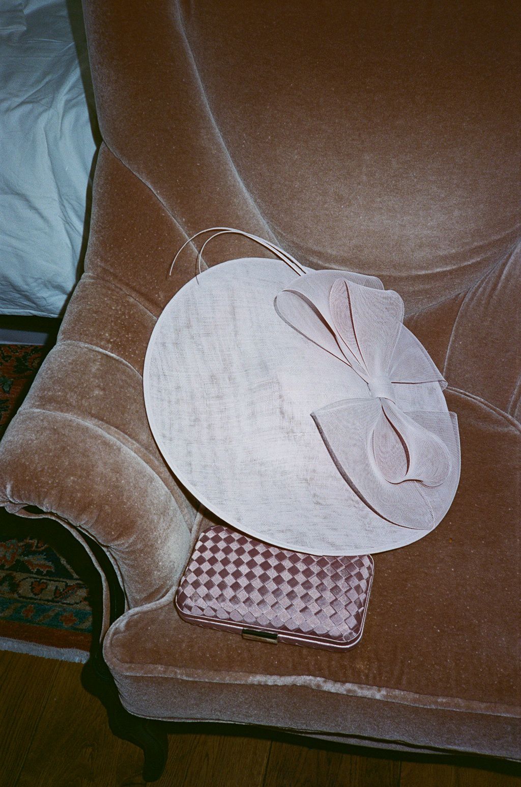A pale pink fascinator hat with bow detail resting on a velvet sofa beside a woven checkered clutch bag.