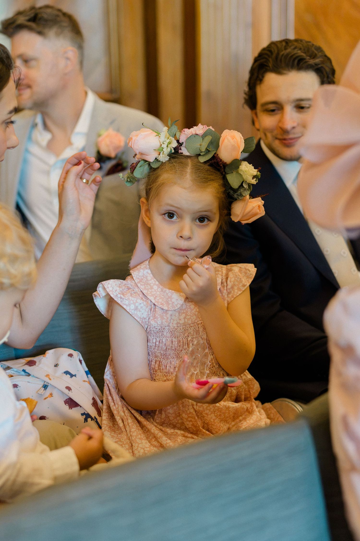 Young flower girl in pink dress and floral crown sits among wedding guests at a ceremony.
