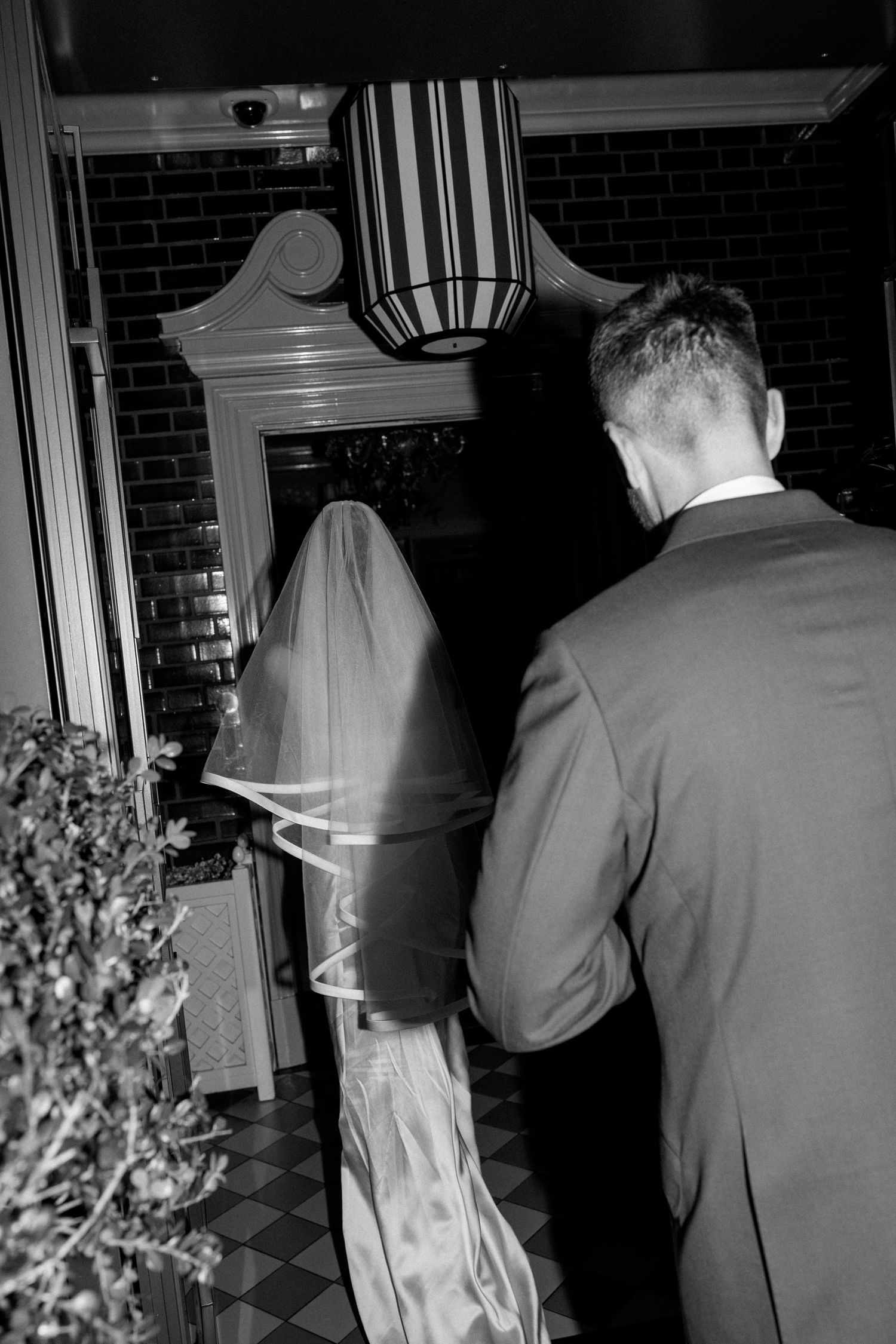 Black and white photo of bride and groom entering elegant venue through ornate doorway beneath striped pendant lamp.