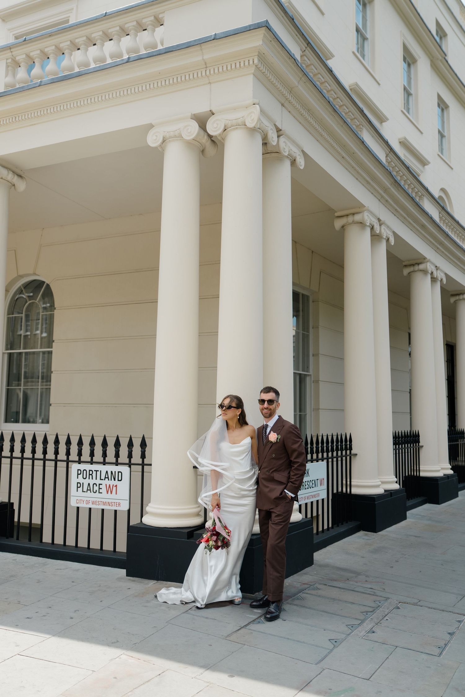 Bride and groom pose outside Portland Place W1 London building with white columns in wedding attire.