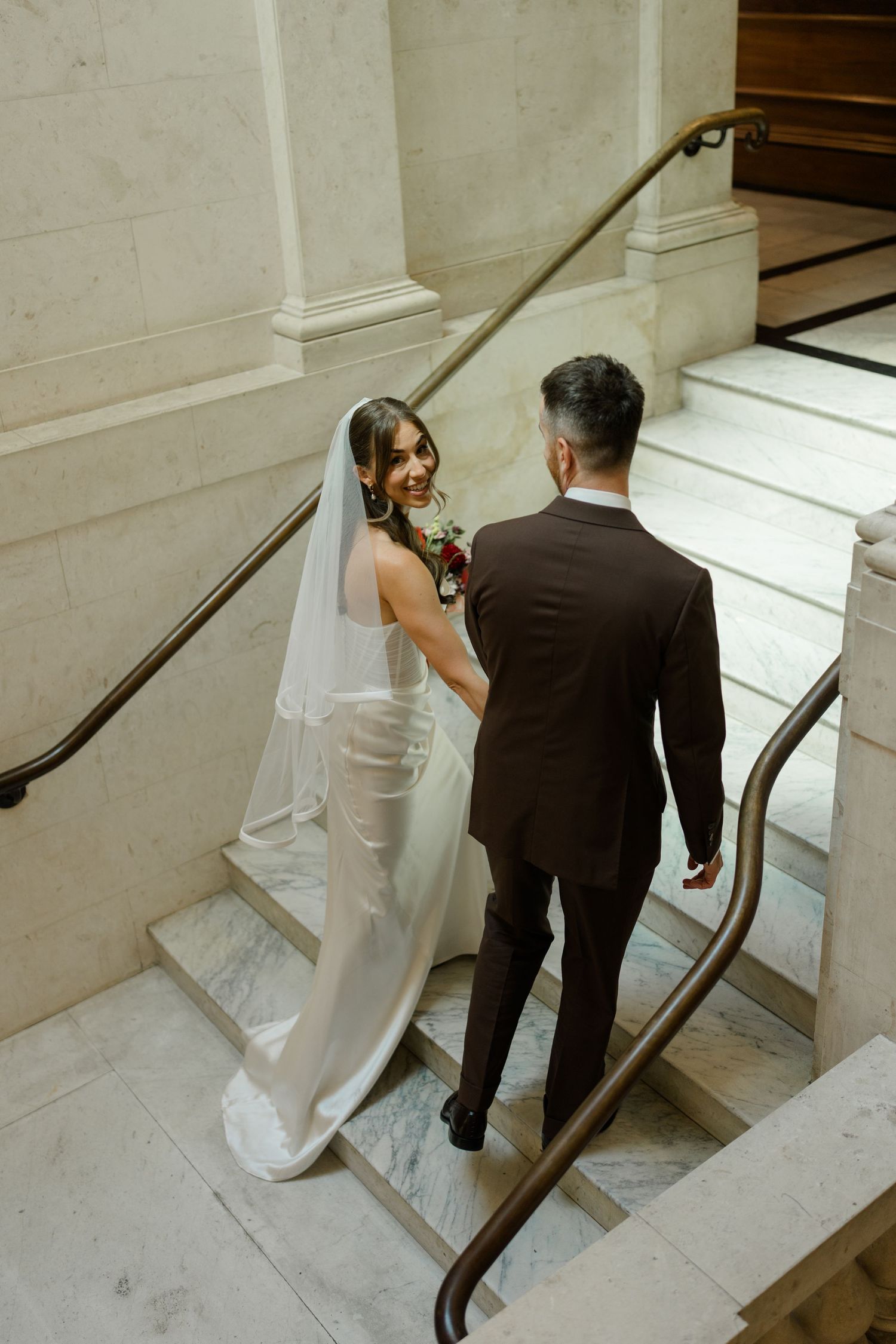 Bride and groom walking up marble staircase, bride in white satin gown with veil, groom in brown suit.