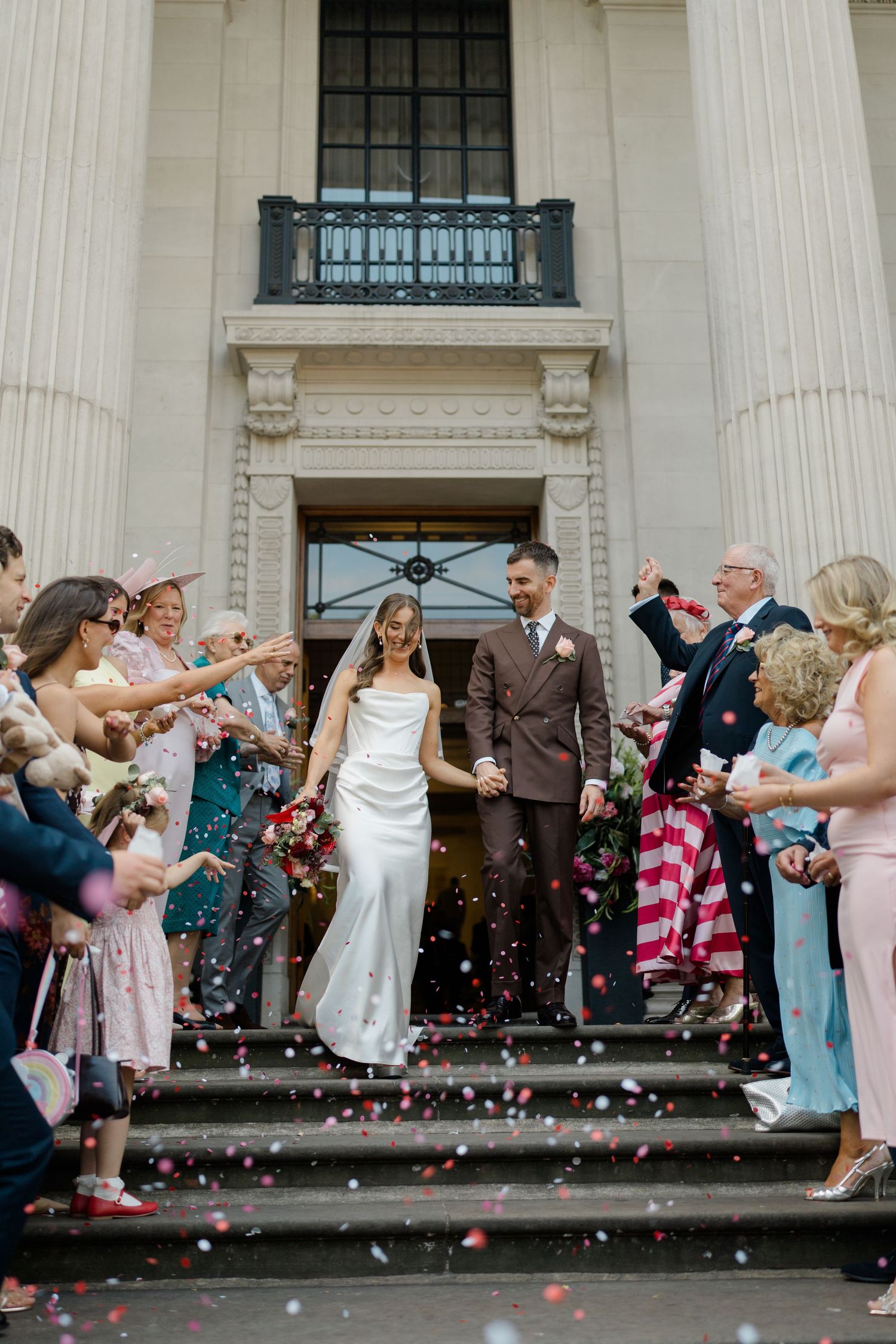 Newlyweds exit neoclassical building steps as guests throw colorful confetti in joyful celebration.