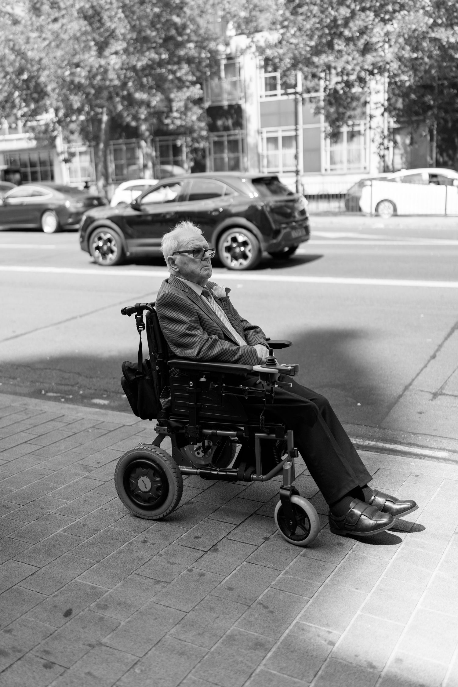Elderly man in suit riding electric wheelchair on city sidewalk in black and white photo.