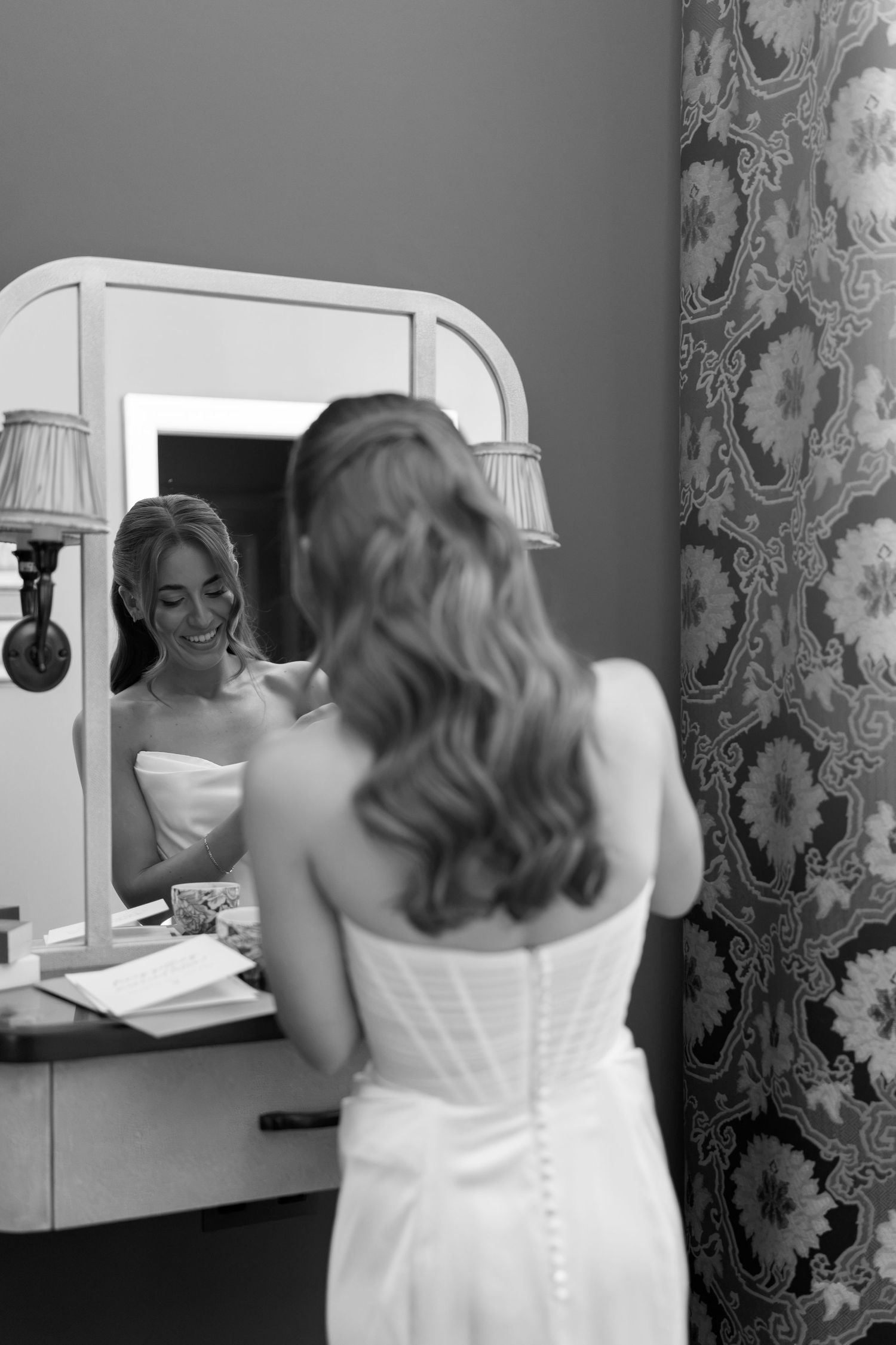 Bride smiling at her reflection in a vanity mirror while getting ready in her white wedding dress.